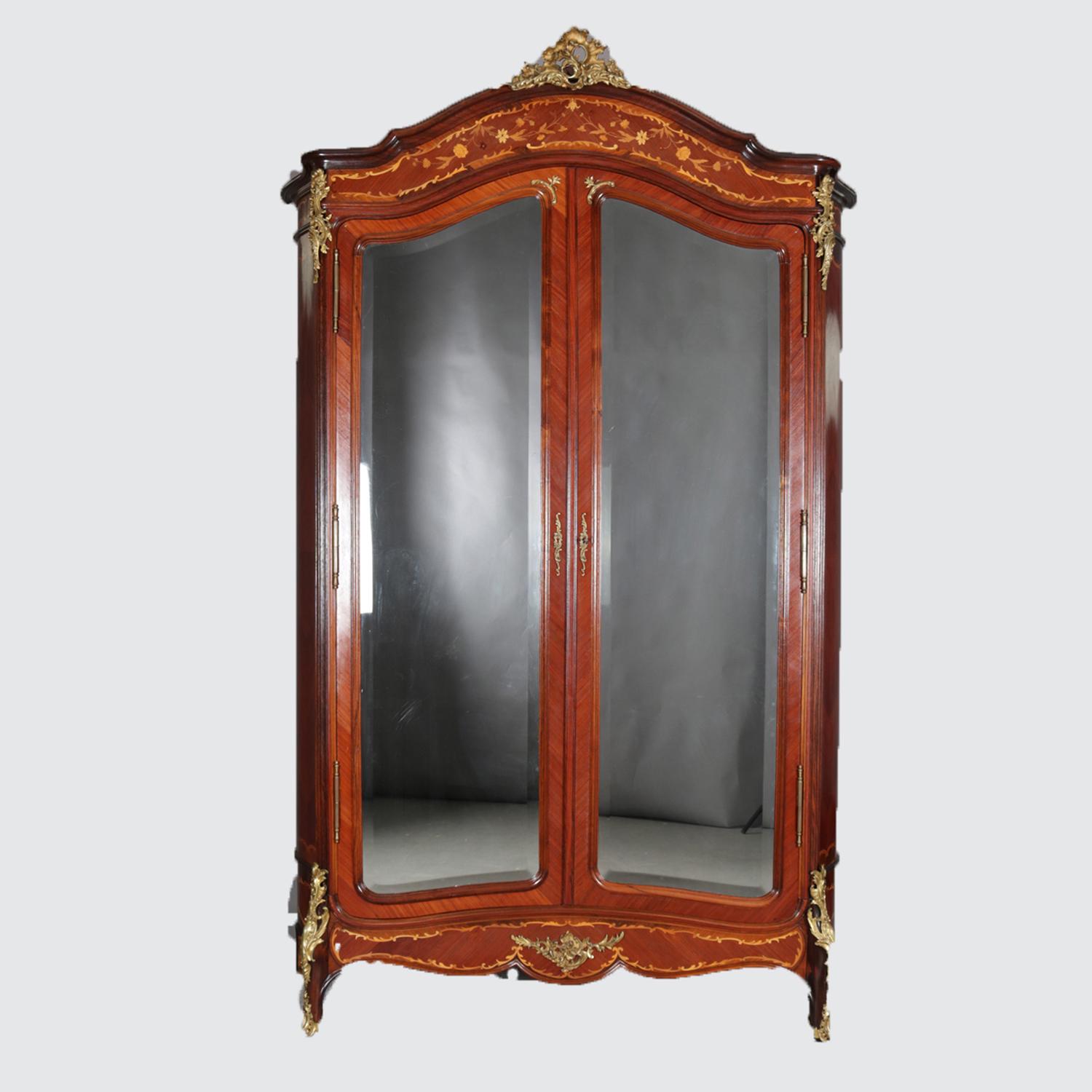 An antique French Louis XIV style mahogany armoire wardrobe features arched crest having foliate form ormolu finial and bookmatched fascia with satinwood inlaid foliate and floral decoration surmounting double doors with beveled mirrors opening to