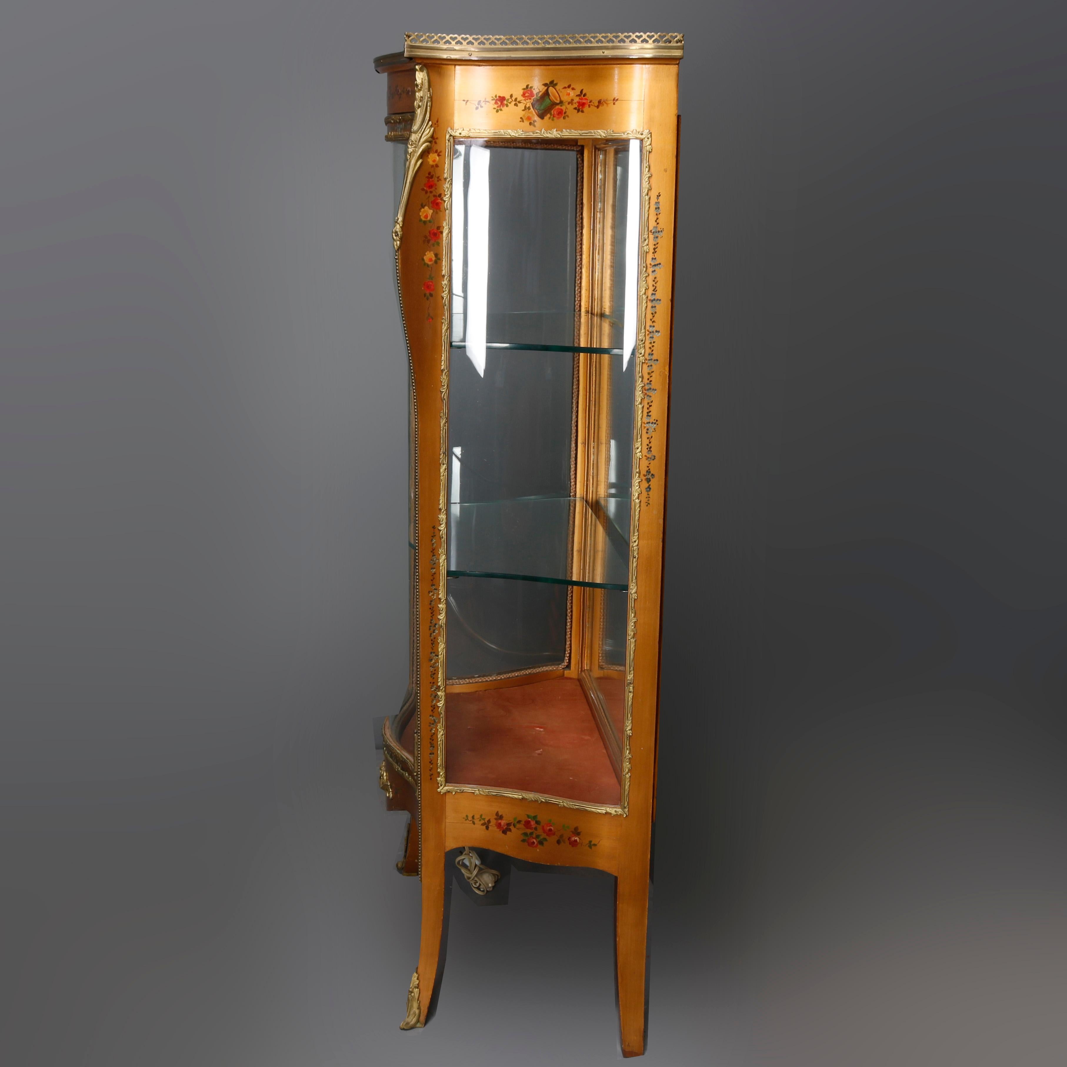 An antique French Louis XIV style vitrine curio cabinet offers pierced bronze gallery surmounting floral paint decorated cherry case having concave bowed glass side panels and convex glass door opening to lighted and mirrored interior with glass