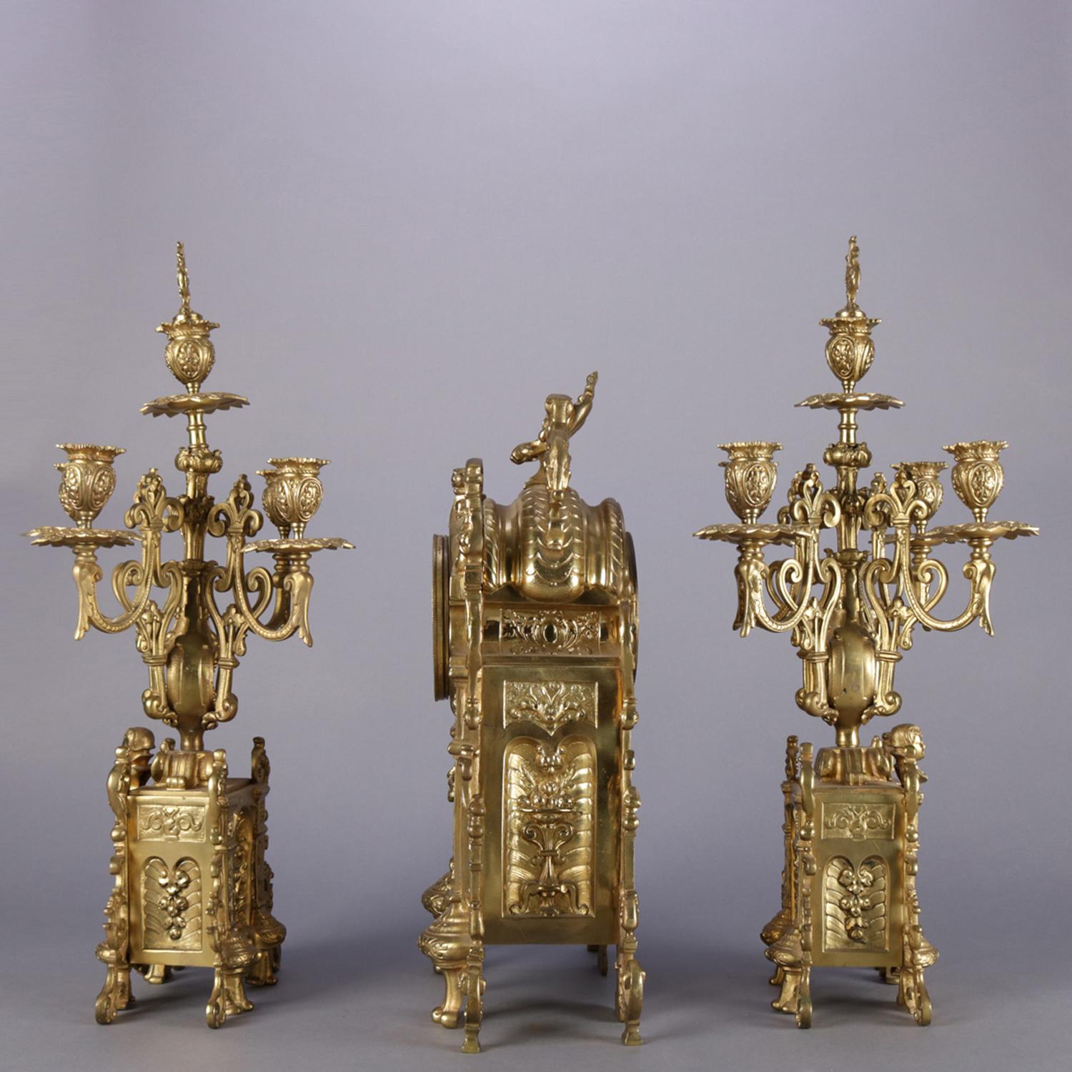 Antique French Louis XV three-piece garniture set features gilt bronze clock with opposing figural lions on crest and case with foliate and scroll decoration and central mask of the king, flaked by matching four arm candelabra with urn and flame