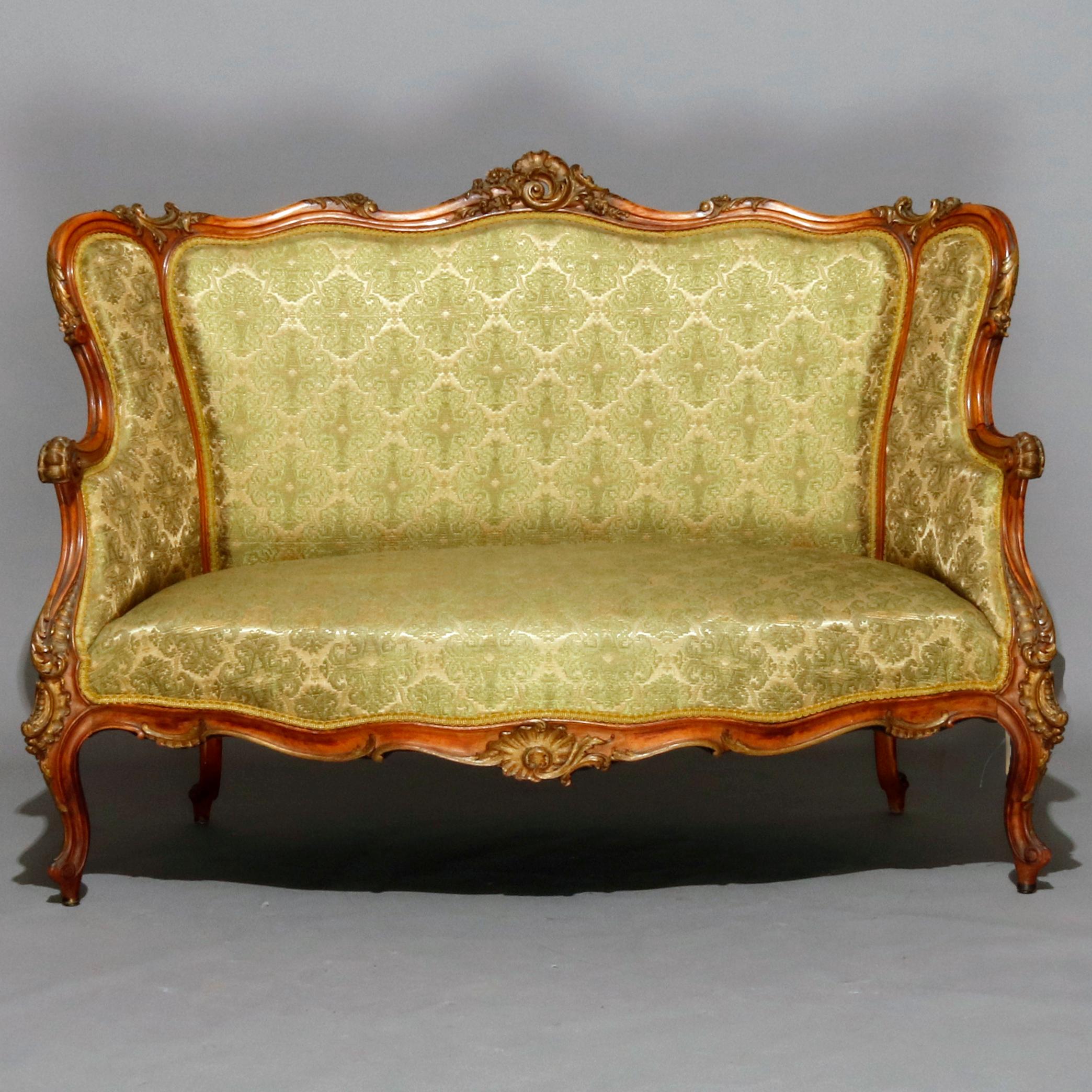 An antique French Louis XV parlor set features wingback form having carved walnut parcel gilt frames with gadroon and foliate crests surmounting shaped backs and scroll form arms, skirt with central carved flower and raised on cabriole legs with