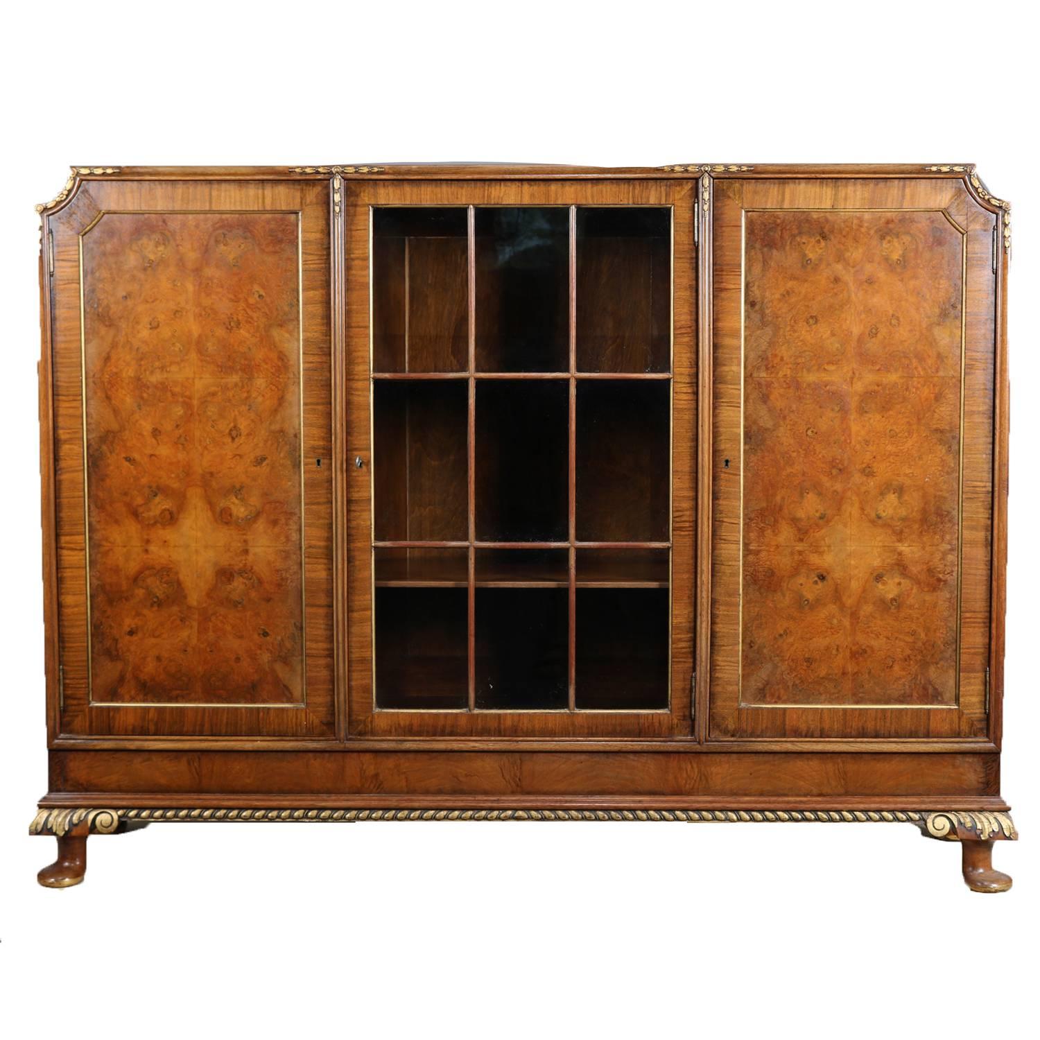 Antique French Louis XV style bookcase features case with upper trim having gilt bellflowers above central glass door flanked by gilt banded doors with burl reserves all opening to reveal three chambers each with adjustable shelving over carved and