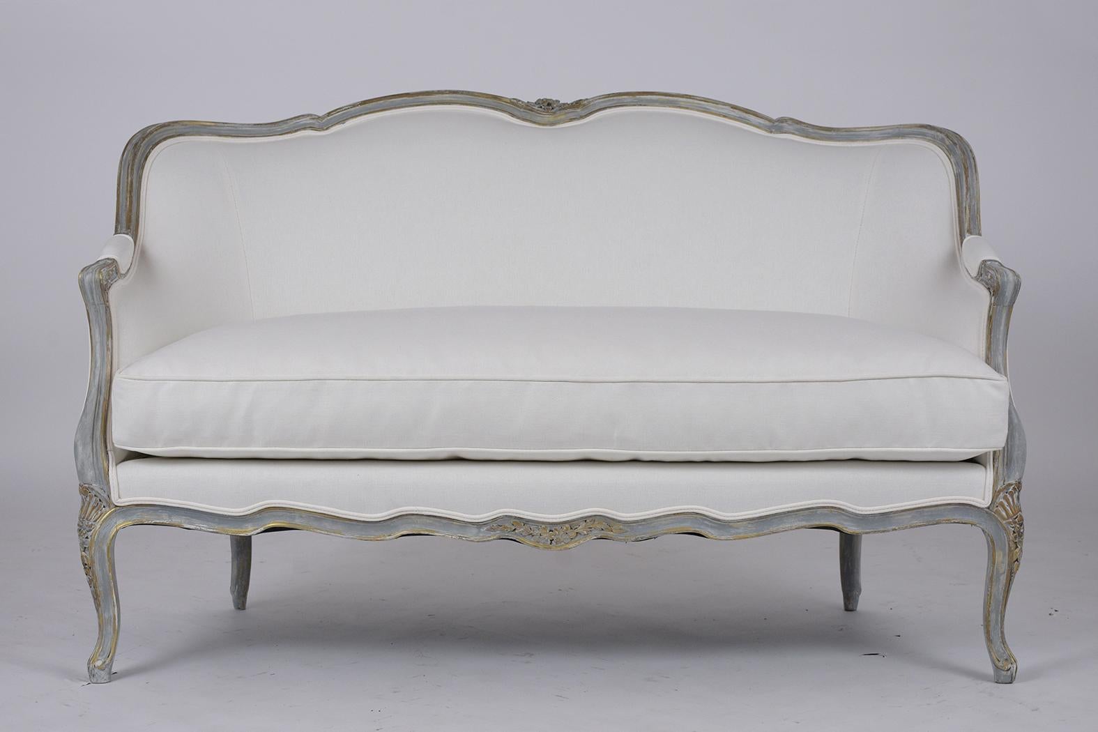 This French Antique Sofa has been newly restored and is made out of walnut wood, and been newly painted in a grey color with gilt accents & distressed finish. The sofa features a curved back with hand-carved floral details on top, scrolled armrests,