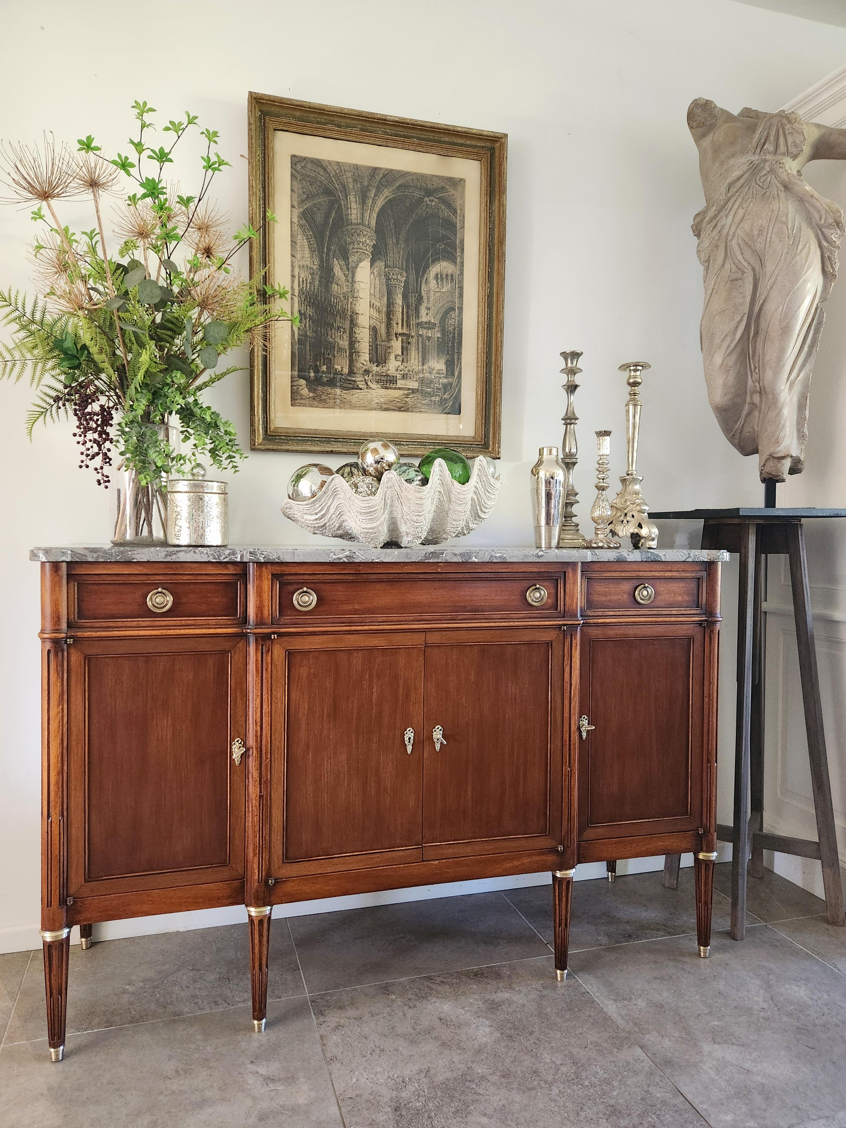 Buffet ancien de style Louis XVI français du 19ème siècle, garni d'un marbre gris, pieds cannelés terminés par des anneaux et sabots en bronze doré.
Trois tiroirs à queue d'aronde et quatre portes et leurs clés, détails en bronze et laiton, marbre