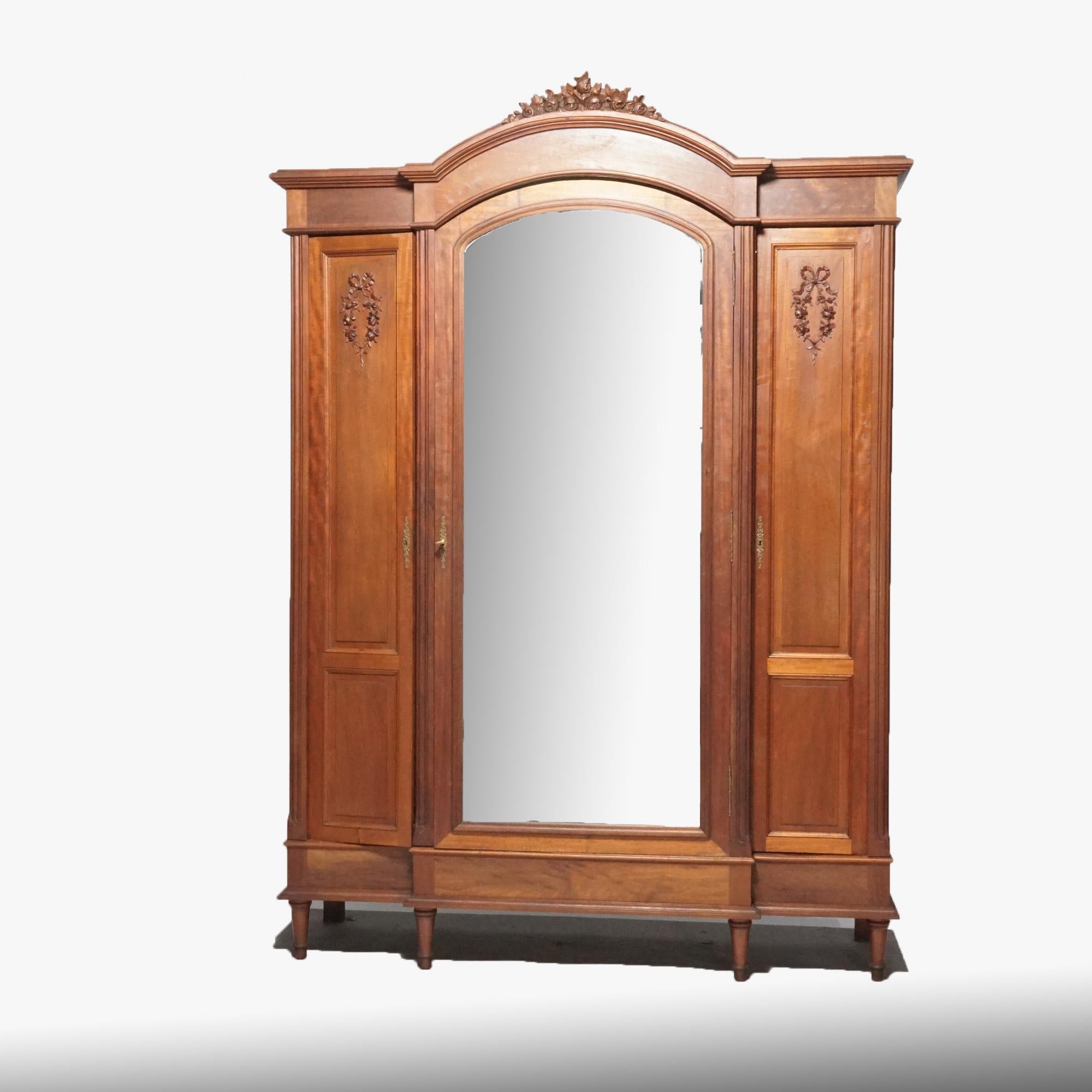 An antique French Louis XVI armoire offers mahogany construction with three cabinets, the central having carved floral crest over arched and mirrored door flanked by two blind doors with carved floral garland decoration, raised on turned and tapered