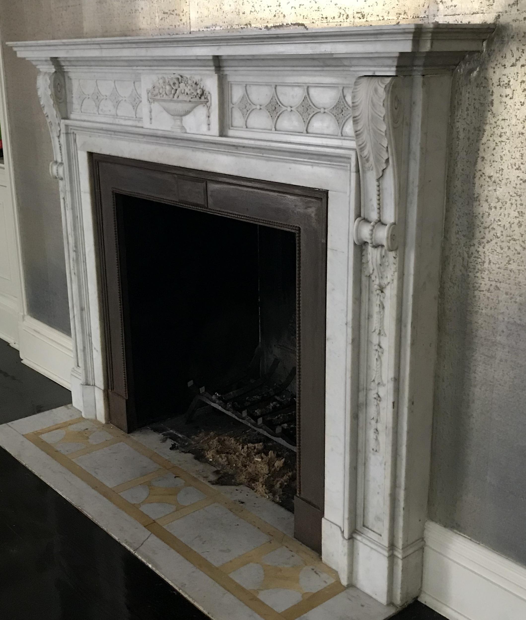 A large Louis XVI style 19th century fireplace surround in Italian Carrara Marble. The carved acanthus leaf console panels flanking each side, terminating in scrolls. The urn frieze with carved flower bouquet in the center, and flanked by a star