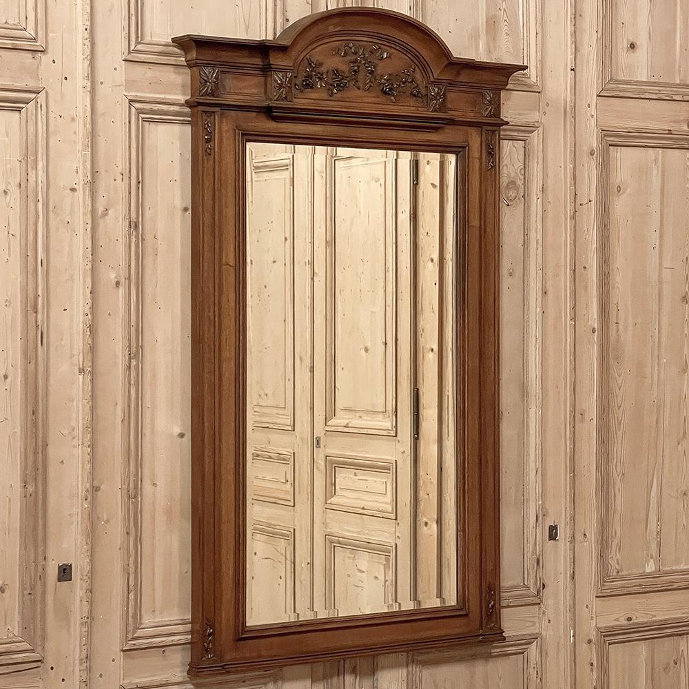 Antique French Louis XVI Carved Walnut Mirror represents the essence of the neoclassical revival style.  The design features a stately architecture enhanced by a boldly molded and arched crown upon which has been carved floral sprays with ribbon in