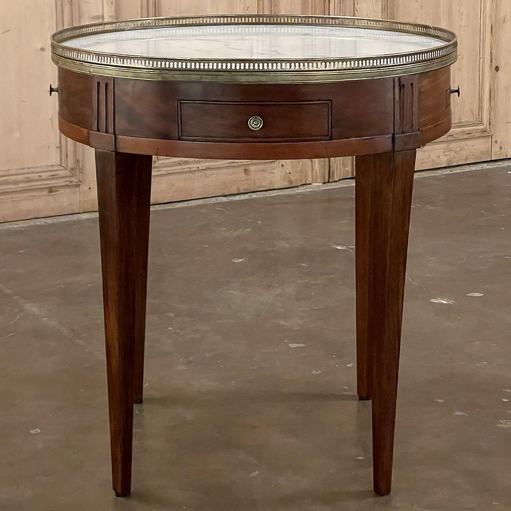 Antique French Louis XVI Mahogany Marble Top Bouillotte Table represents an understated elegance that is perfect for the tailored yet refined decor.  Hand-crafted from exotic imported mahogany, it features a round apron interrupted by fluted plinth