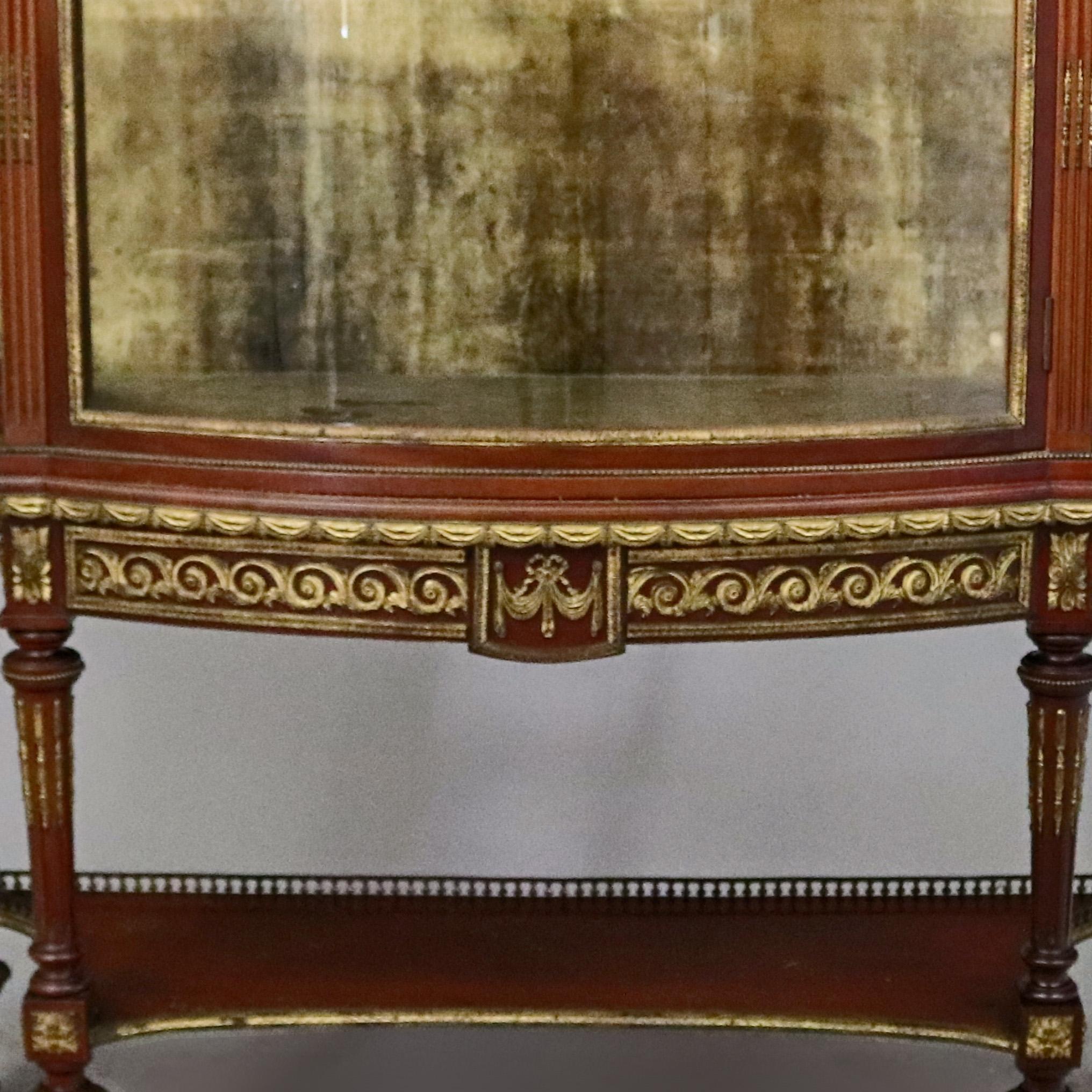 An antique French Louis XVI vitrine features mahogany construction with pierced gallery over case with convex glass sides and single concave glass door opening to glass shelved interior, skirt with cast drape and foliate ormolu mounts, raised on