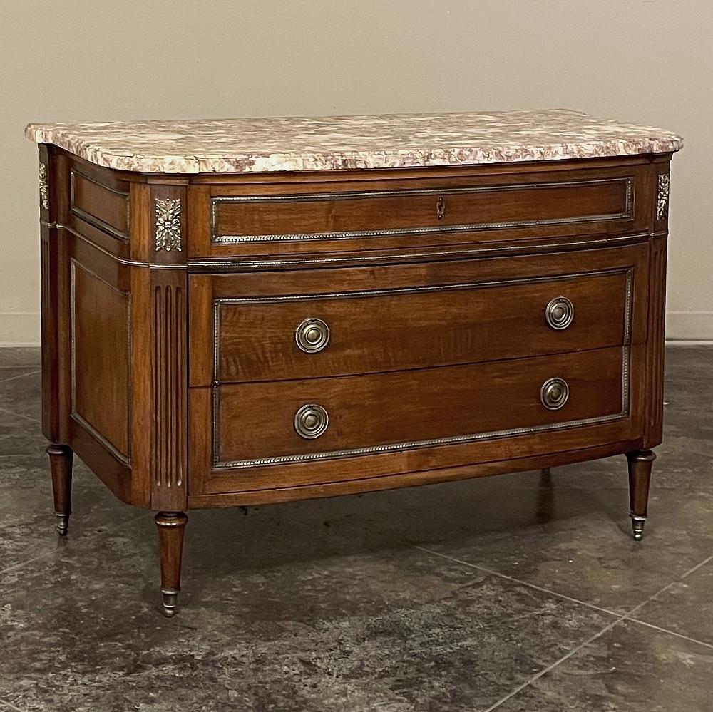Antique French Louis XVI marble top walnut commode was hand-crafted in the neoclassical style and features truly timeless architecture. The gracefully and subtly bowed front blends into rounded, fluted corners which flare at the back against the