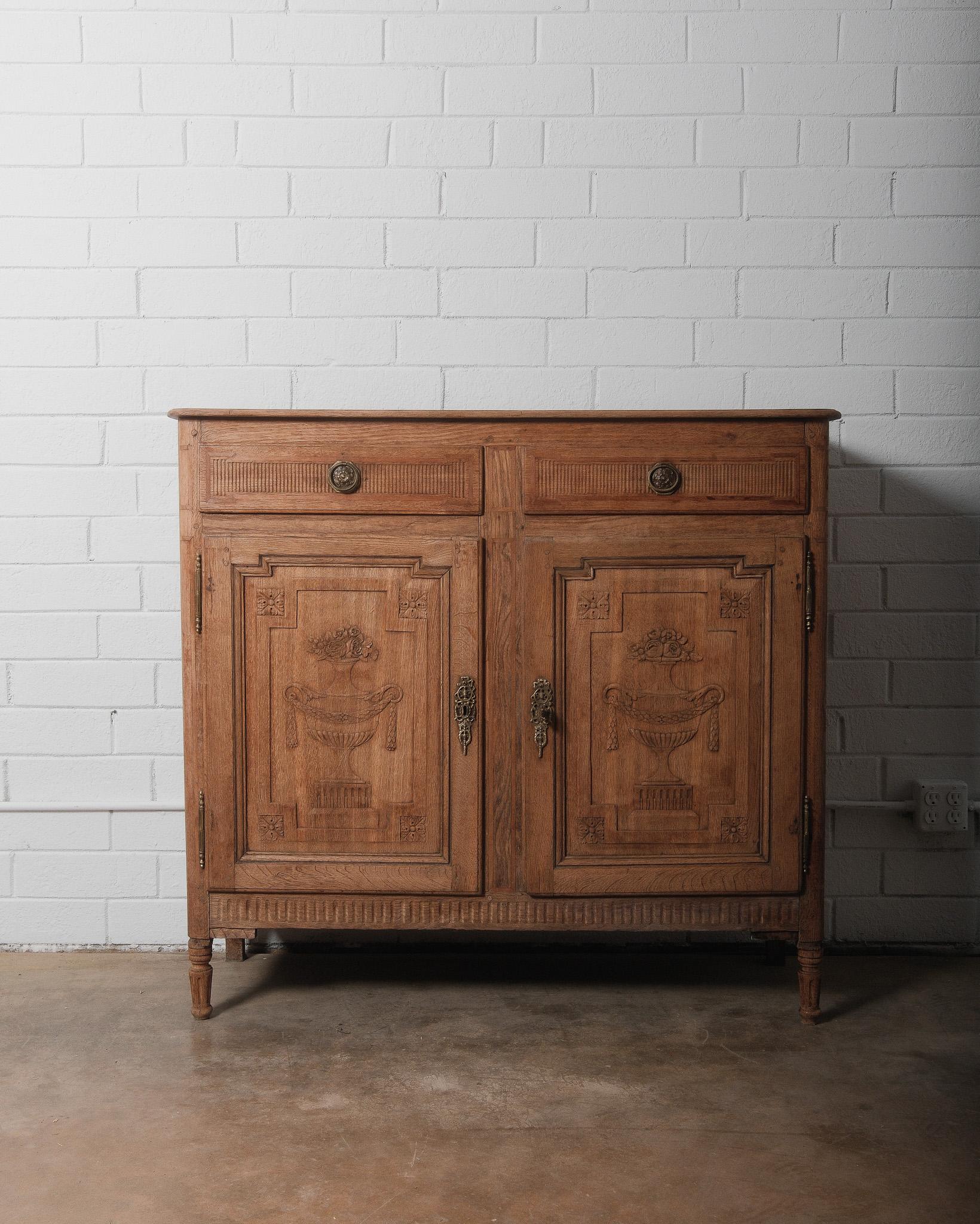 This Louis XVI oak buffet is a stunning example of late 18th-century French craftsmanship. The front doors feature hand-carved vase motifs, framed by intricate geometric patterns and floral detailing, embodying the elegance of the Louis XVI era.