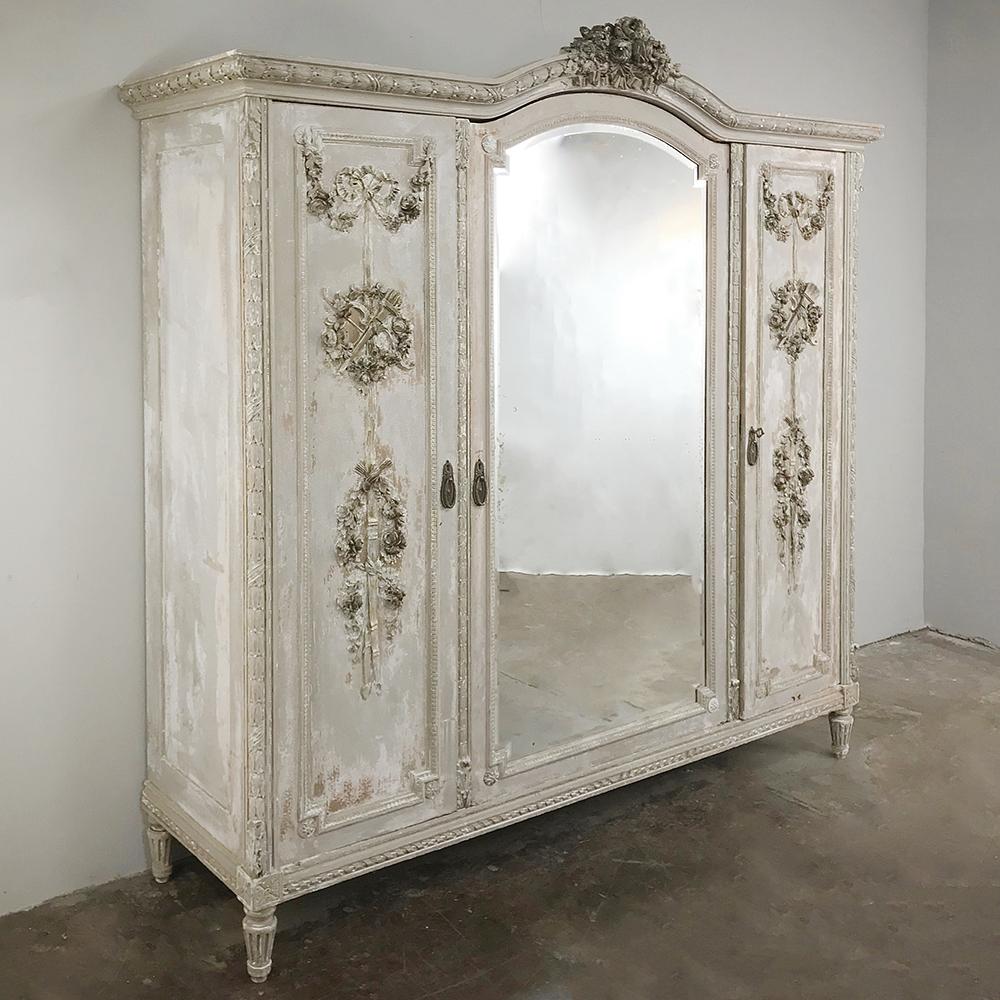 Antique French Louis XVI Painted Armoire features a full length mirror on the center door, with fine carved detail on the side doors depicting the quiver of arrows and bow with floral wreaths and swags, enhanced by the two-toned distressed painted