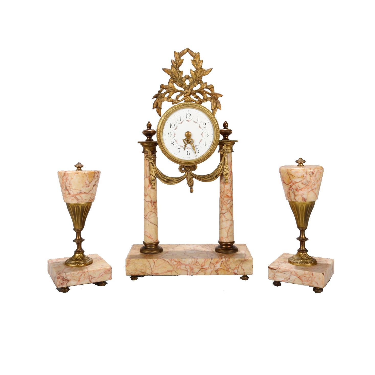 An antique French Louis XVI garniture set offers central clock having foliate cast bronze crest over works having flanking rouge marble column supports with cast bronze draped swag and two complementing rouge marble stylized torchieres, bronze
