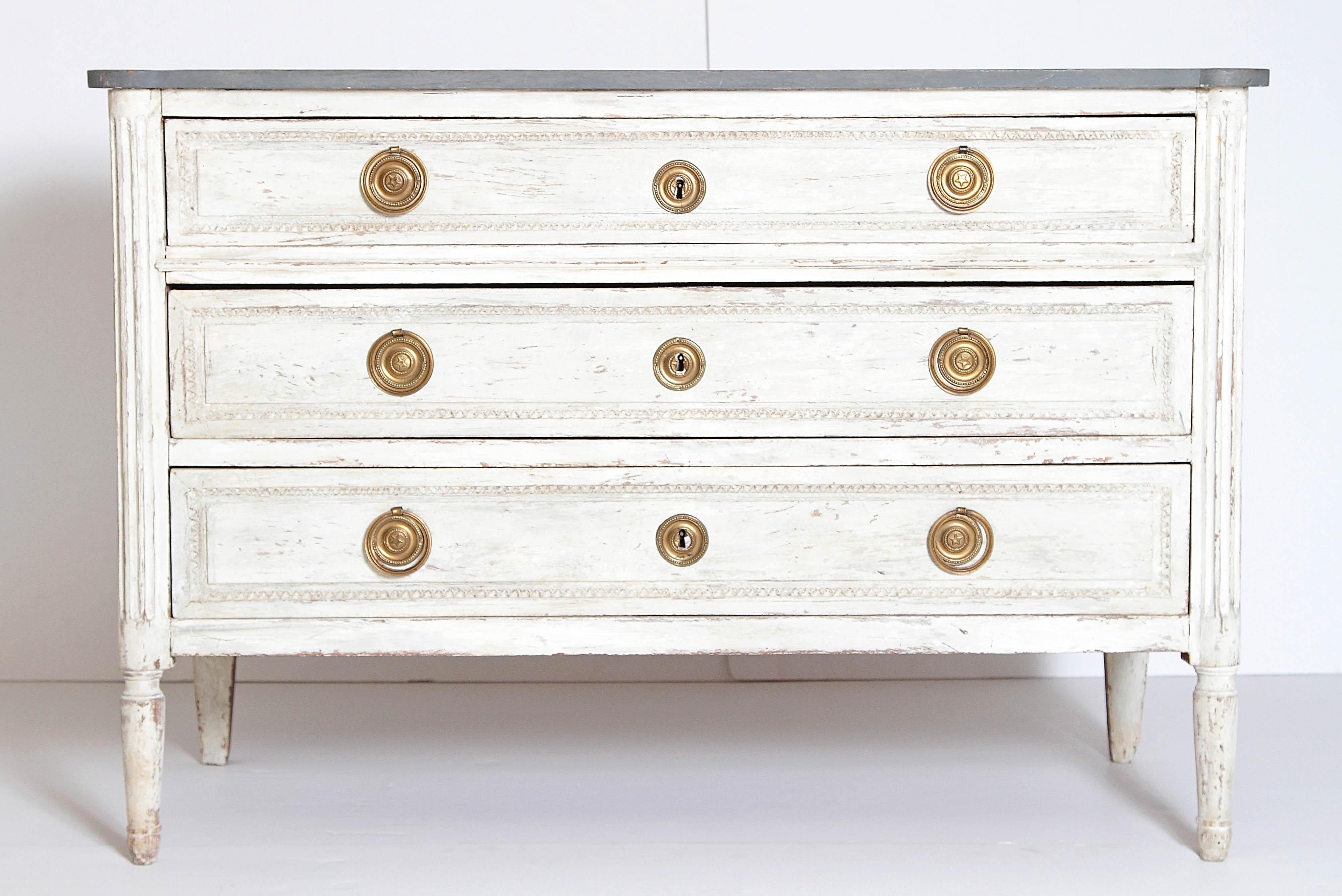 An 19th century antique Swedish Louis XVI style chest of drawers topped with a painted grey wood top. Fluted columns frame the three drawers with carvings. The fluted columns terminate in round fluted legs. Three drawers with round metal pulls with