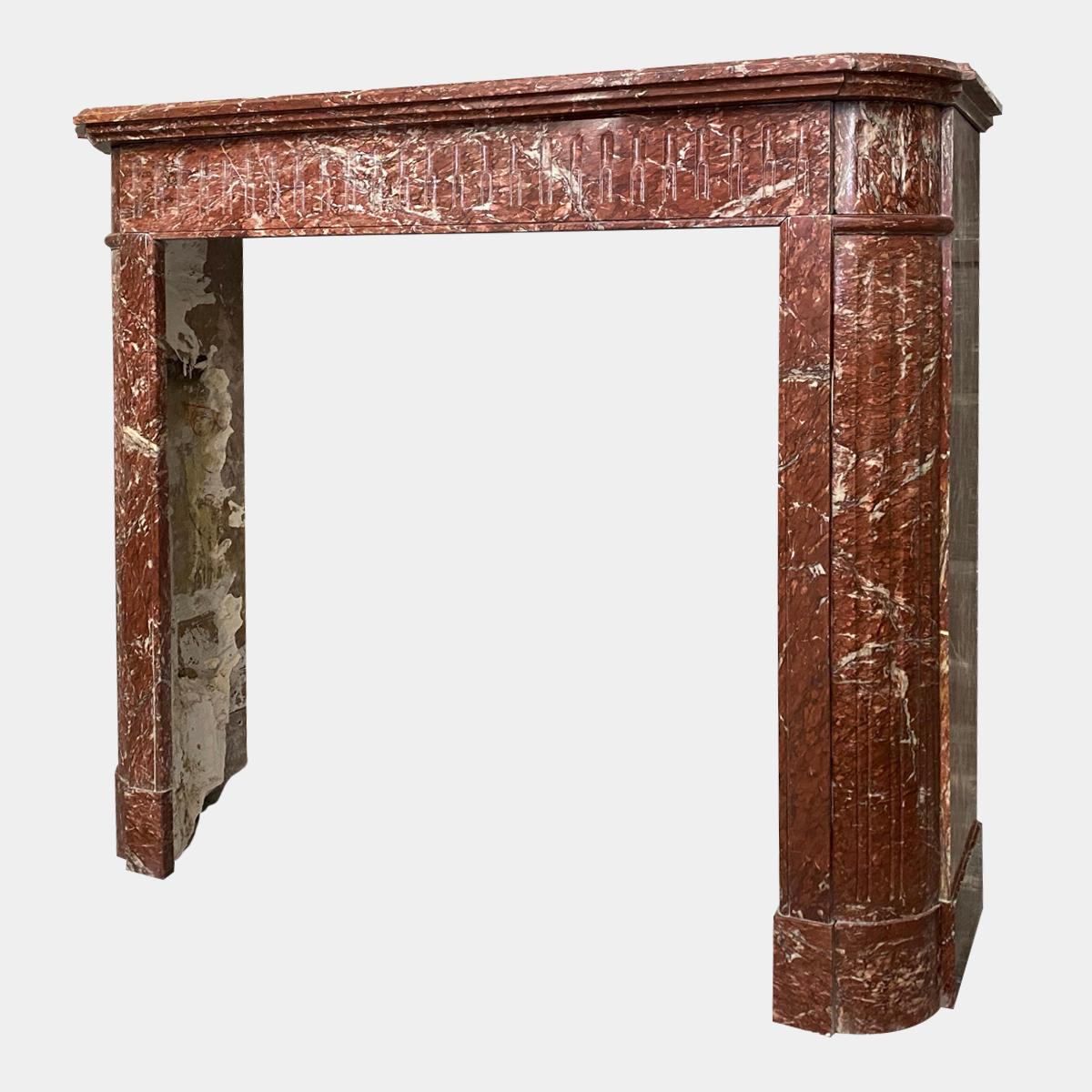 A 19th century petit Louis XVI style fireplace in quality Rouge de Saint Pons marble. The canted jambs with stop flutes panels surmounted by conforming stop fluted corner blocks, flanking a running stop fluted frieze. All beneath a stepped and