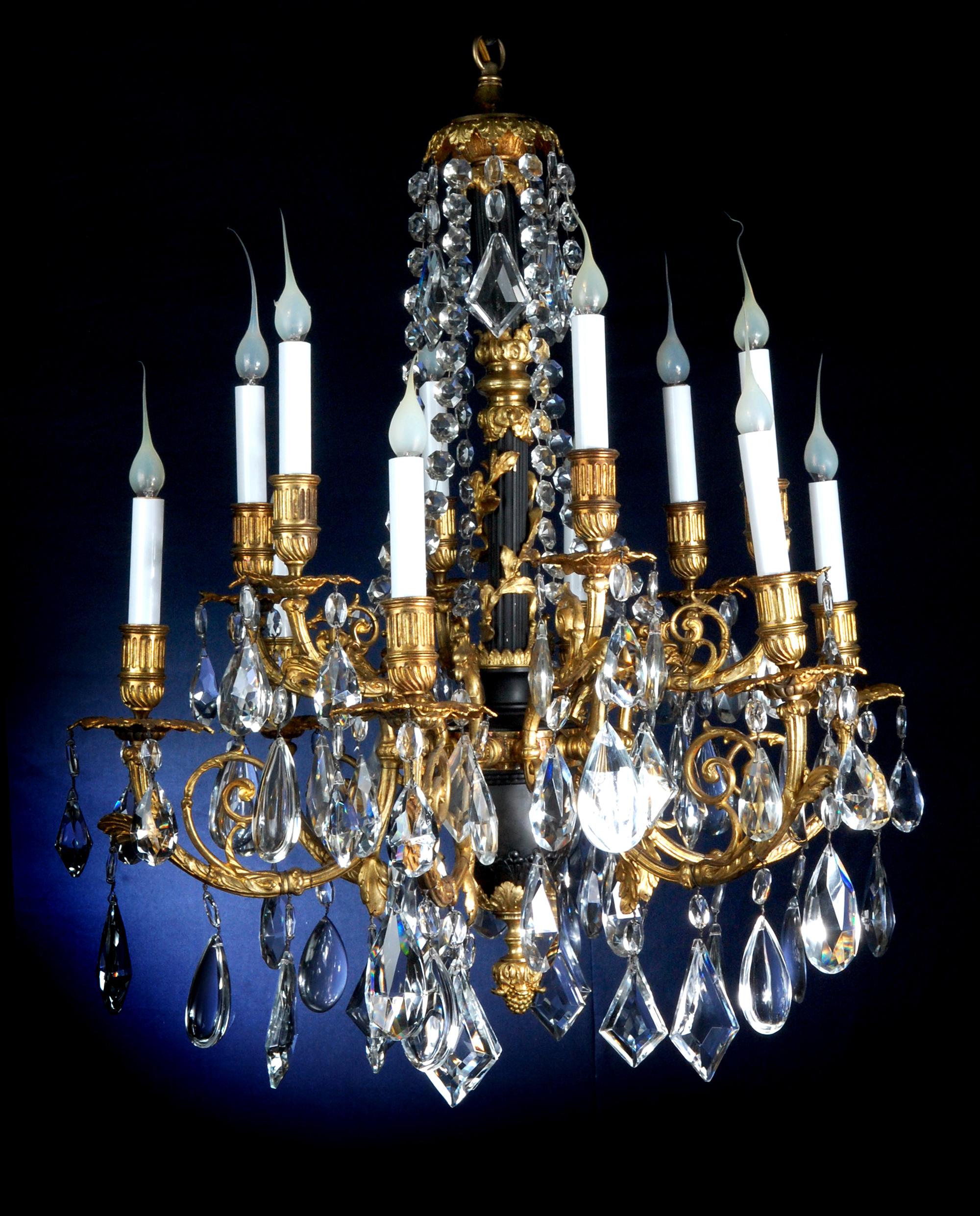 Ce lustre de style Louis XVI, finement moulé, présente un arrangement majestueux de douze lampes en forme de bougie montées sur deux niveaux gradués. Les bras en bronze doré s'enroulent vers l'extérieur, chacun se terminant par une buse cannelée et