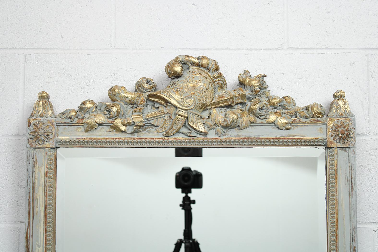 This French antique Louis XVI style wall mirror is in good condition and has been newly painted in a grey and white color combination with a distressed finish. The mirror features remarkable carved details along the top depicting a decorative