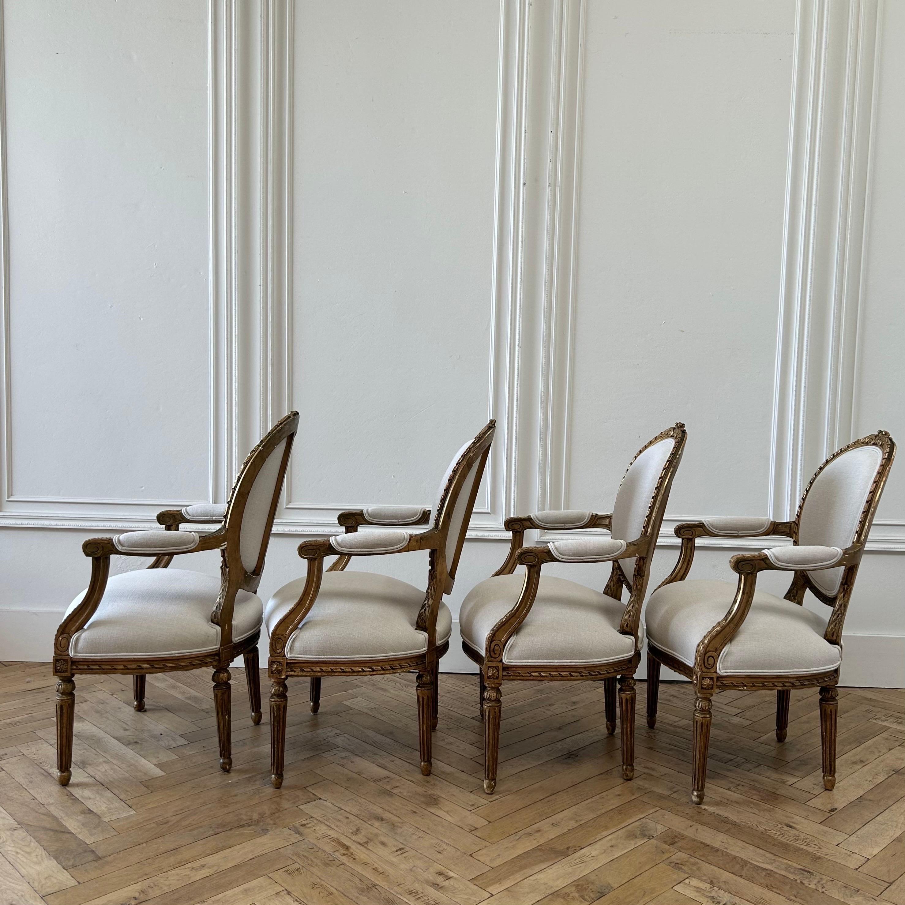 4 beautiful French Louis XVI Style Open arm chairs in original gilt wood finish.
Distressed finish, solid and sturdy ready for daily use.
New upholstery in Libeco oatmeal linen.
Size: 24”w x 24”d x 37”h. 
SH:19”. SD:19”. AH:25”
Sold per Chair.