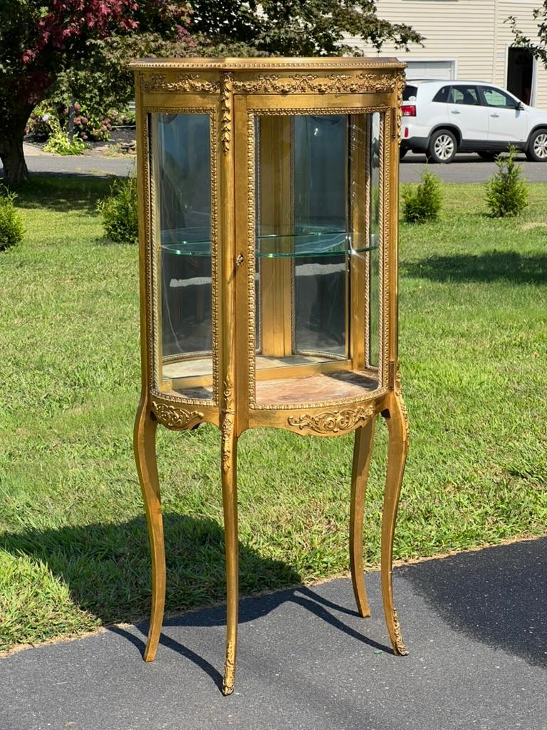 Cette exquise vitrine en bois doré de style Louis XVI, réalisée par l'estimé Geo. C. Flint Co. de New York, respire l'élégance et le goût raffiné caractéristiques du début du XXe siècle. Le délicat cadre doré de la vitrine, orné de motifs finement