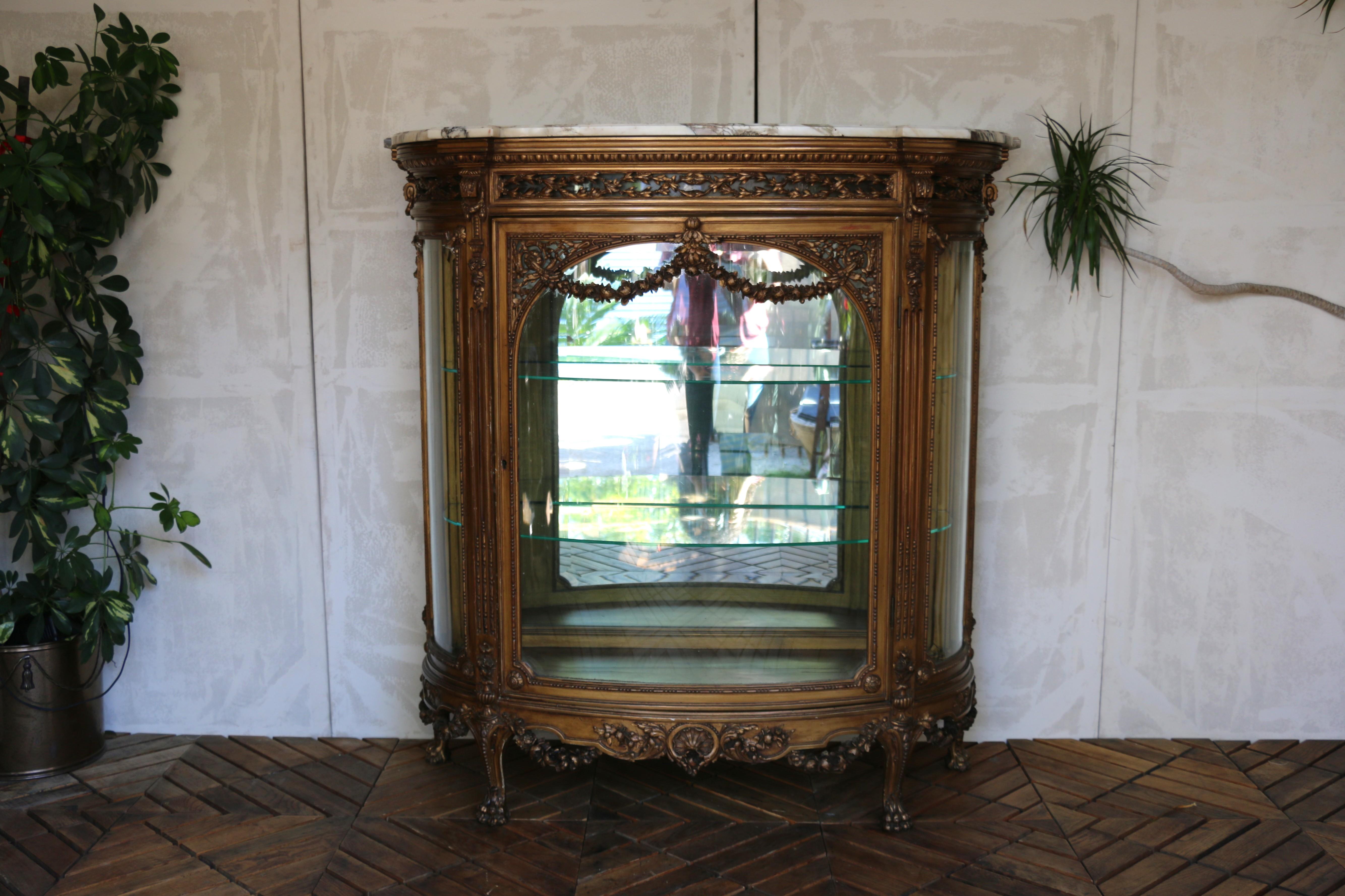 Esta exquisita vitrina francesa antigua muestra los opulentos detalles y la artesanía característicos del estilo Luis XVI. El mueble presenta un marco de madera dorada ricamente tallada, adornado con guirnaldas florales, motivos de cintas, pedrería