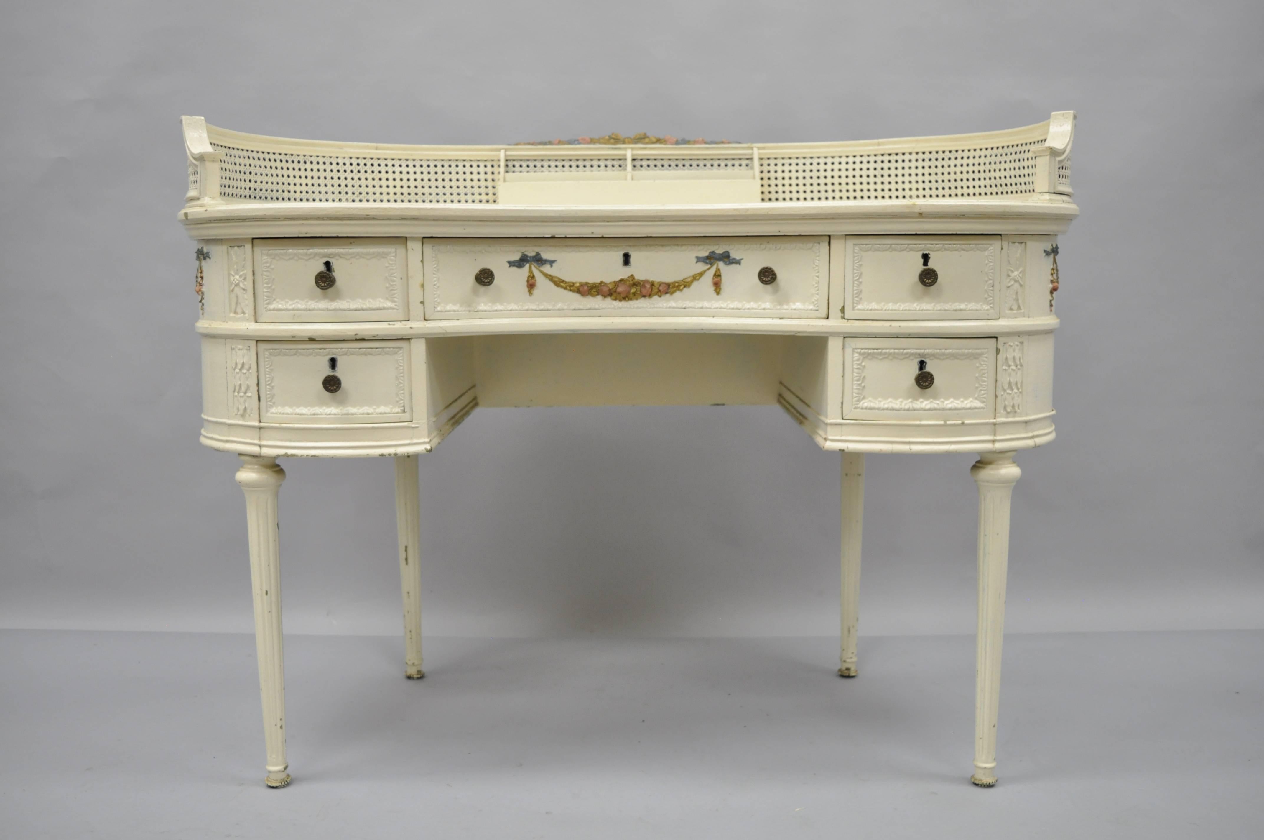 Antique French Louis XVI style kidney shaped writing desk or vanity table. Item features a unique kidney shape, finished back, floral drape applied carvings to drawer front and sides, cane gallery with storage slots, distressed white painted finish,
