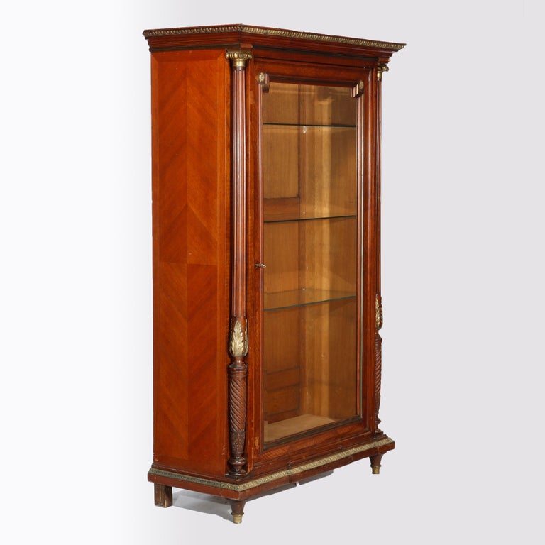 An antique French Louis XVI style cabinet offers kingwood construction with bookmatched facing, single glass door opening to shelved interior and having flanking Corinthian column supports, raised on Mersailles bun feet, foliate cast ormolu