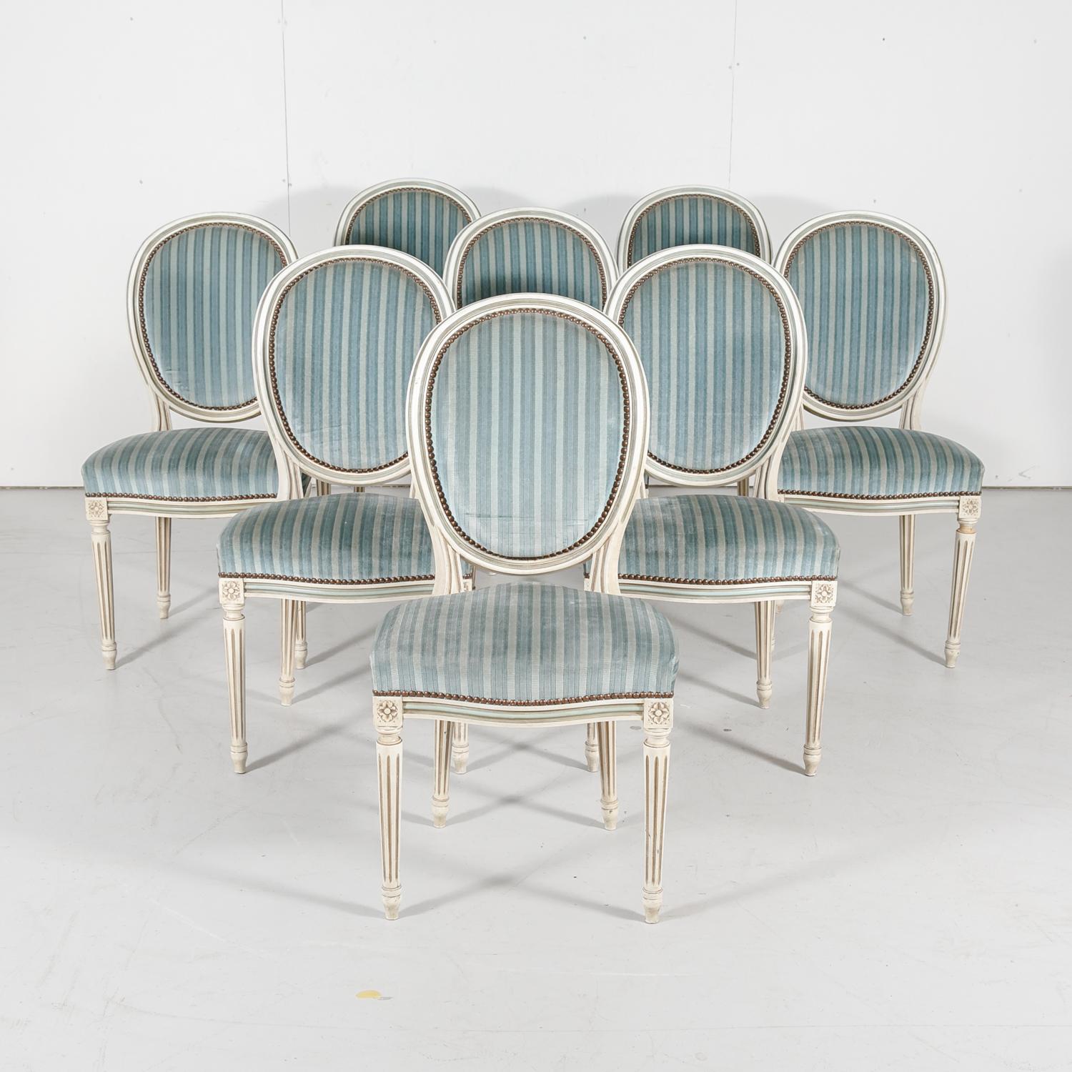 Set of eight French Louis XVI style carved and painted side chairs having the original cream painted finish with accents of French blue and taupe paint, circa 1920s. Upholstered medallion shaped backs and serpentine seats with nail head trim, all