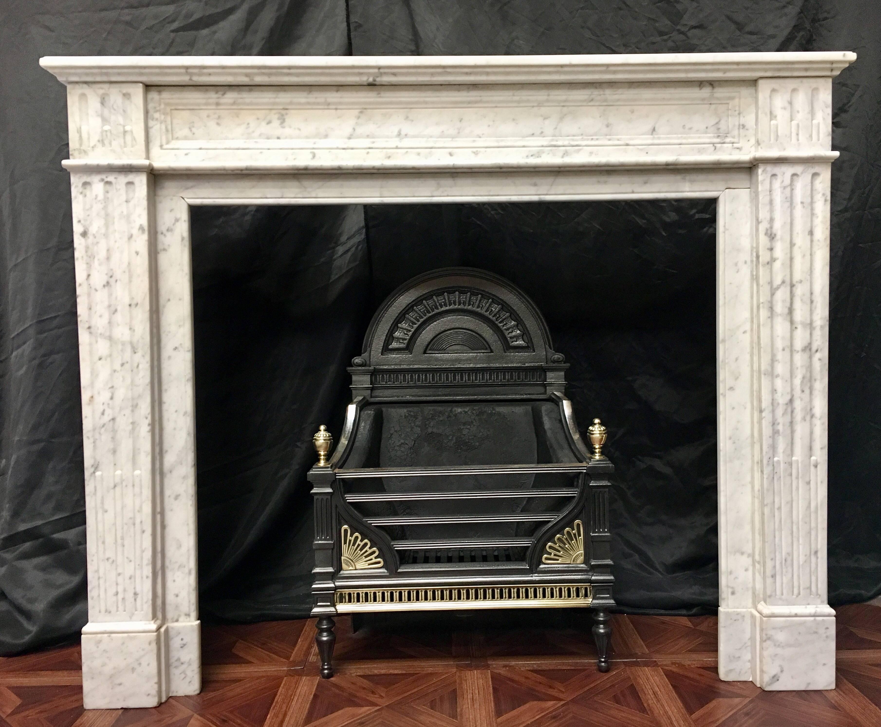 An antique 19th century French Louis XVI style white Carrara marble fireplace surround, of simple form. A moulded shelf sits above a recessed and moulded frieze, flanked by capitols of fluting supported by tapered empire style jambs with stop