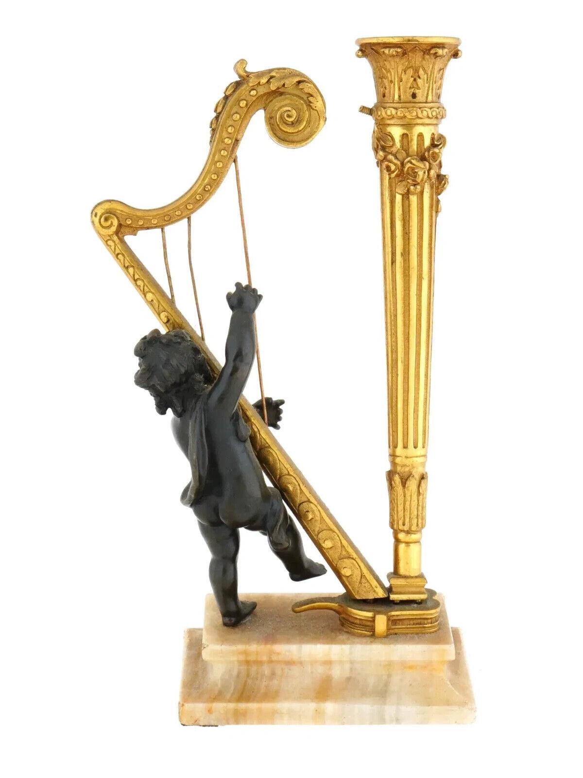 Antigua lámpara escultórica de bronce patinado dorado a la francesa. La lámpara tiene un diseño neoclásico que representa a un querubín tocando un arpa. La base de la lámpara está colocada sobre un pedestal cuadrado de mármol de color pálido. Sin