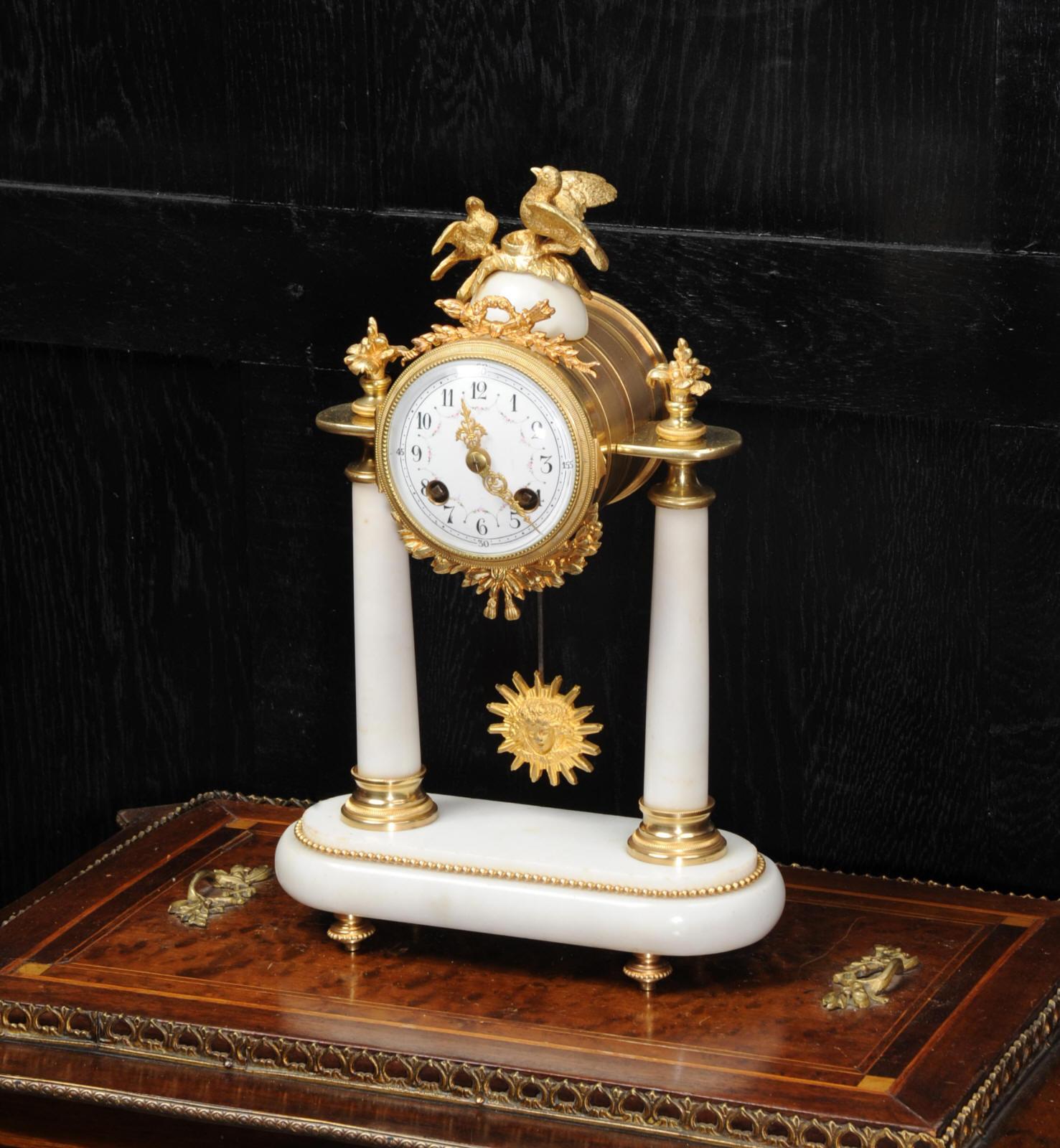 A beautiful original antique French portico clock of white marble mounted with ormolu (finely gilded bronze). The movement is held aloft on two marble pillars with the ormolu pendulum swinging gently below. Above is a pair of nesting birds,