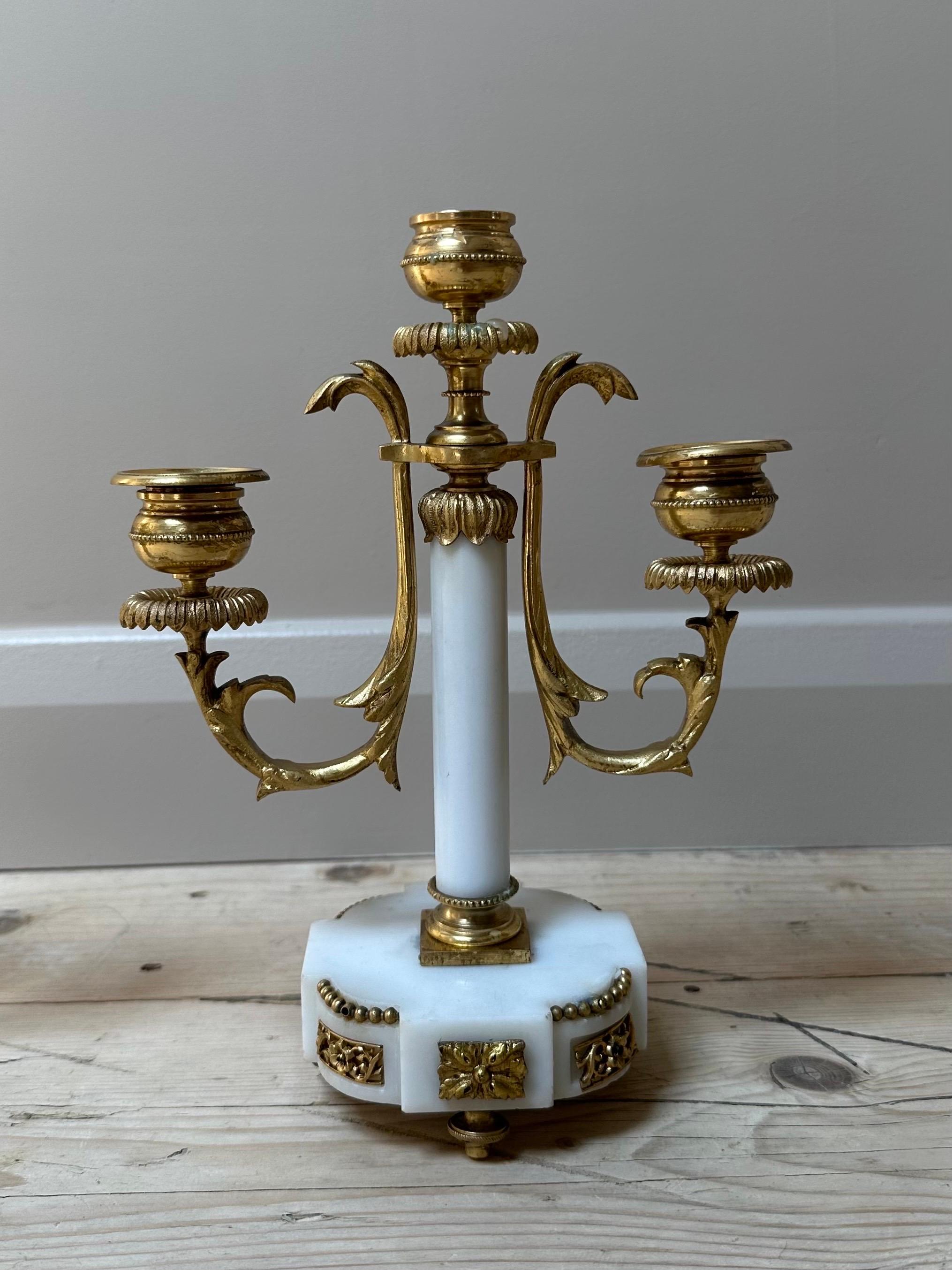 Cette exquise paire de candélabres français anciens est magistralement fabriquée en marbre blanc et en laiton doré orné. Chaque candélabre présente trois bras gracieusement arqués, ornés de motifs classiques de feuilles d'acanthe, s'étendant à