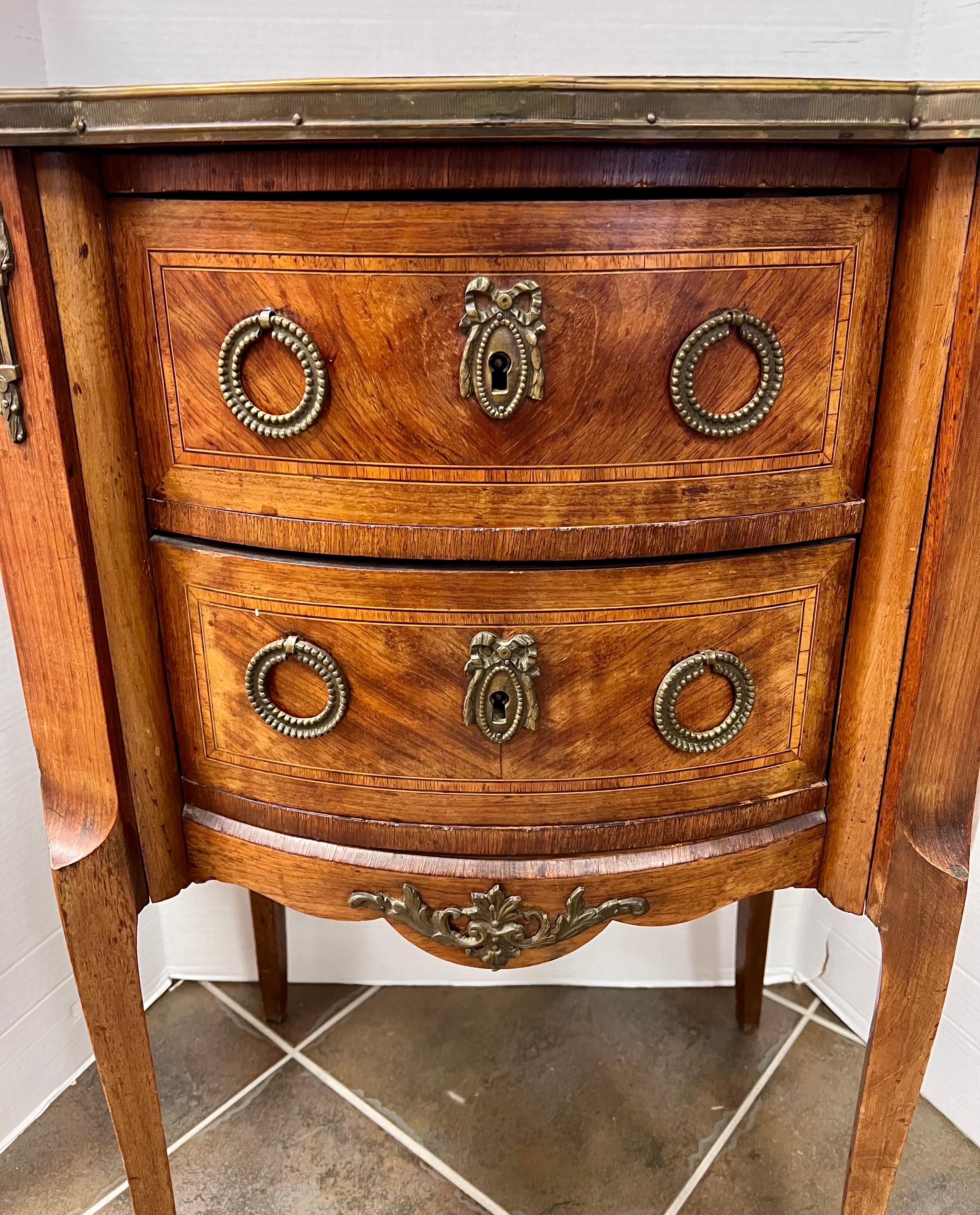 French Louis XV style commode nightstand features inlaid detail and pink marble top with brass gallery. Two drawers, cabriole legs, and mounted ormolu detailing.