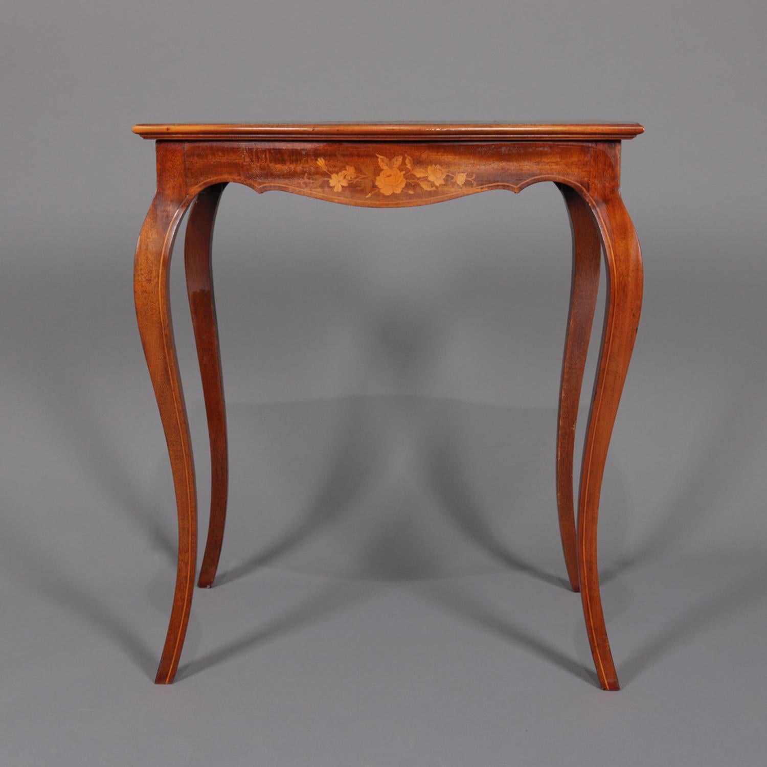 An antique French marquetry lamp table features mahogany construction with shaped top having central inlaid floral bouquet surmounting floral and foliate inlaid scalloped skirt and raised on cabriole legs, satinwood banding throughout, circa