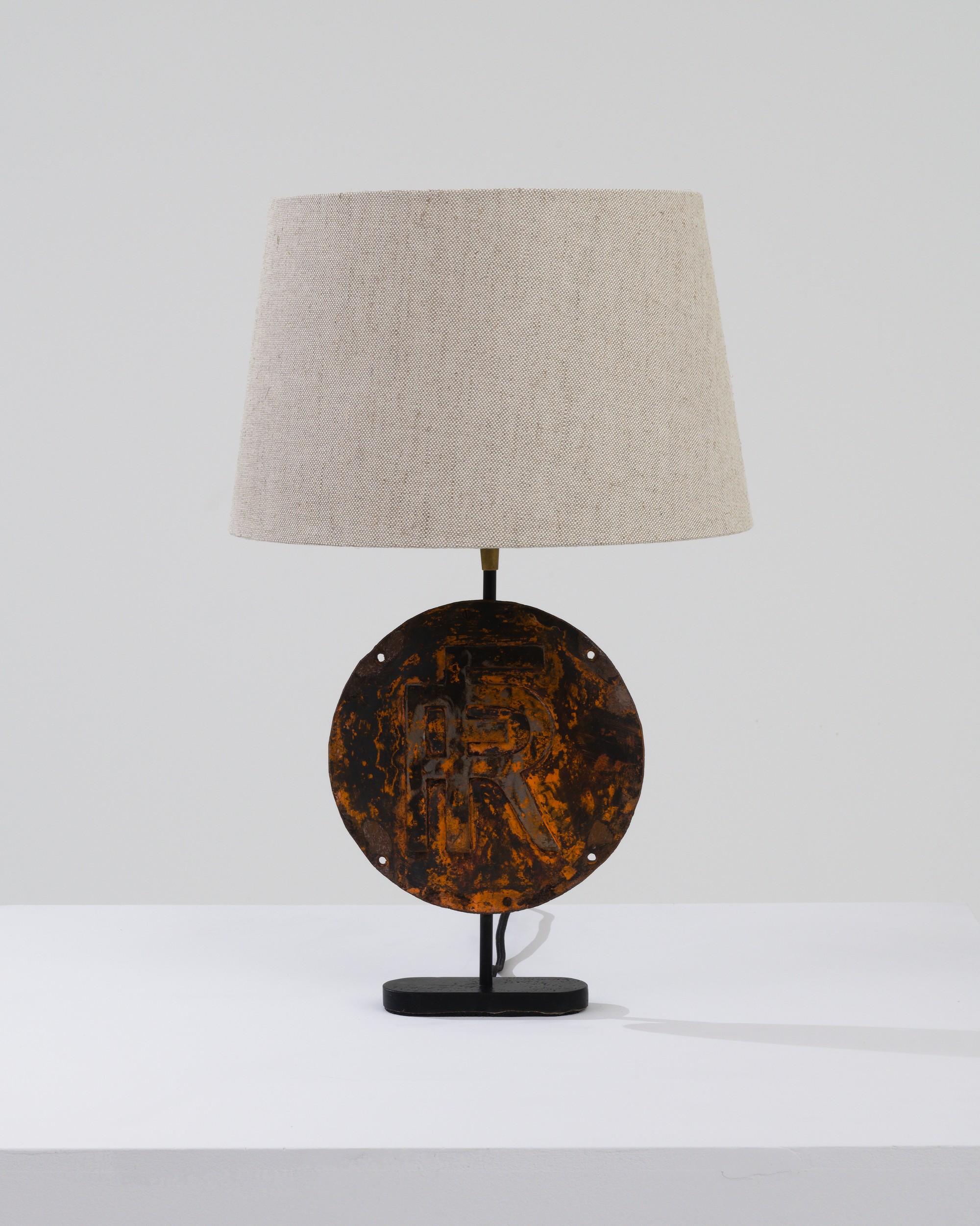 This antique table lamp was crafted in France around the 1900s. The metal base, featuring a circular plate with initials intricately merged, serves as a captivating focal point of this piece. The orange rust on the metal creates a striking marbled