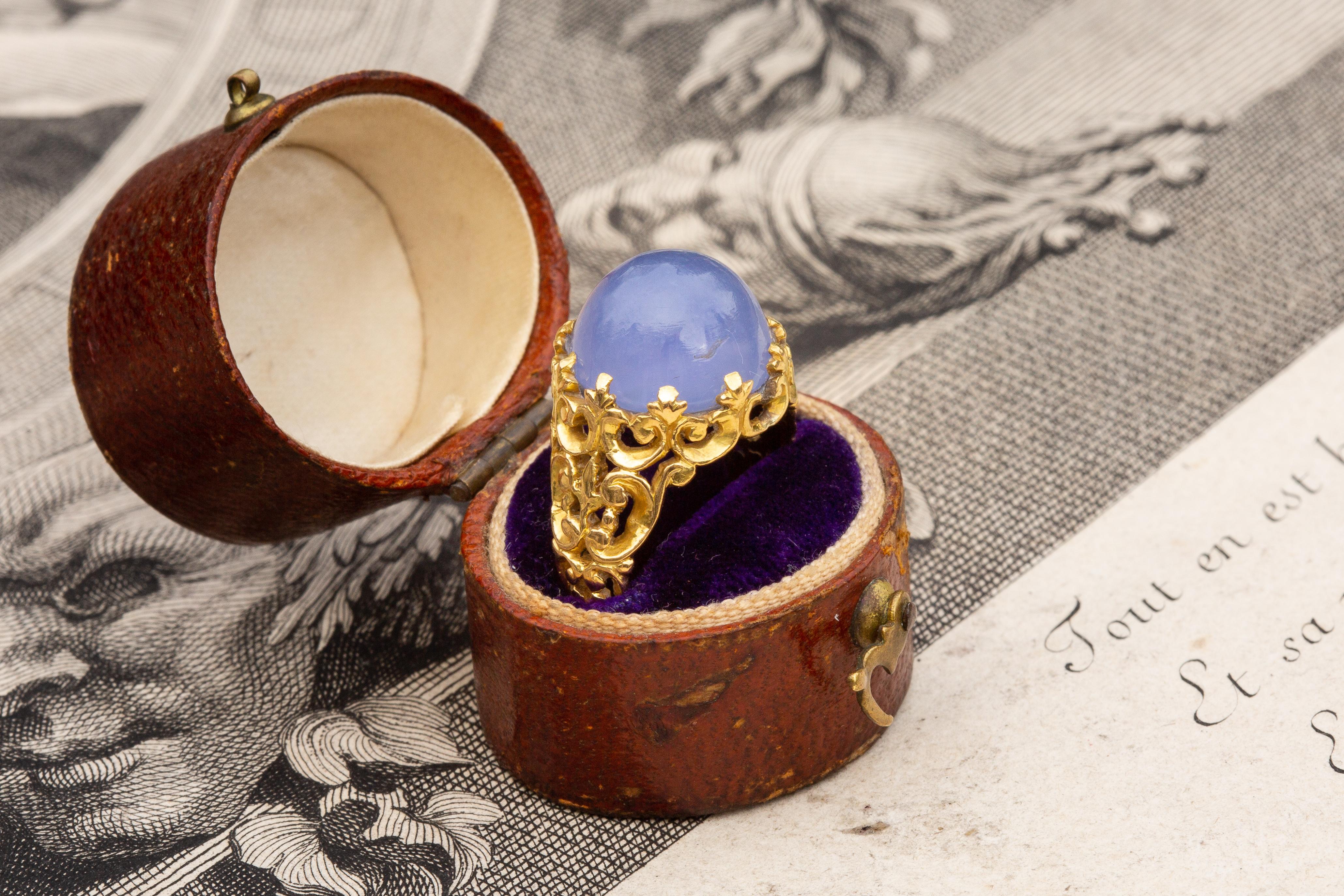 This stunning piece was made in France and dates to the mid-19th century. The ring is crafted in 18K gold and set with a large domed 9.5ct lilac blue chalcedony cabochon in a pronged setting. The blue agate stone rests in an incredibly intricate