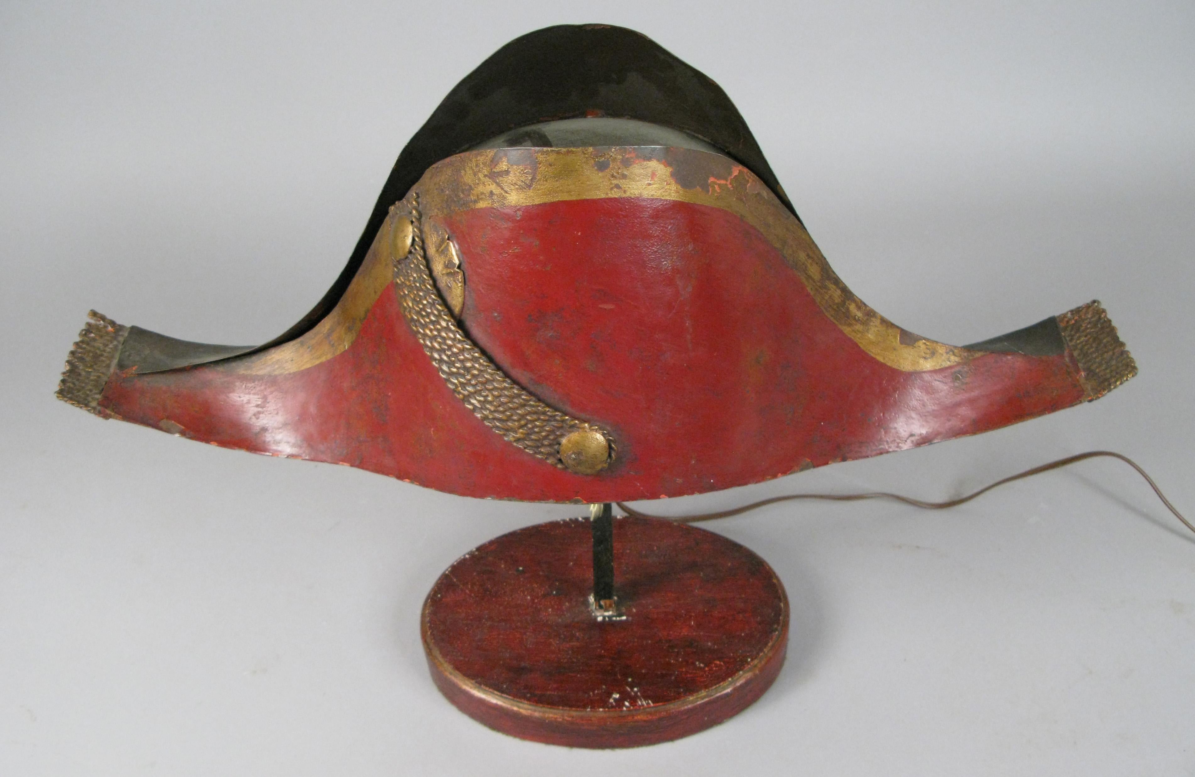 A very charming early 20th century lamp in the form of a tole metal Napoleonic chapeau, mounted on a wood base. Wired to accept a small bulb with a standard base. It is really more of a lighted object than a light source.