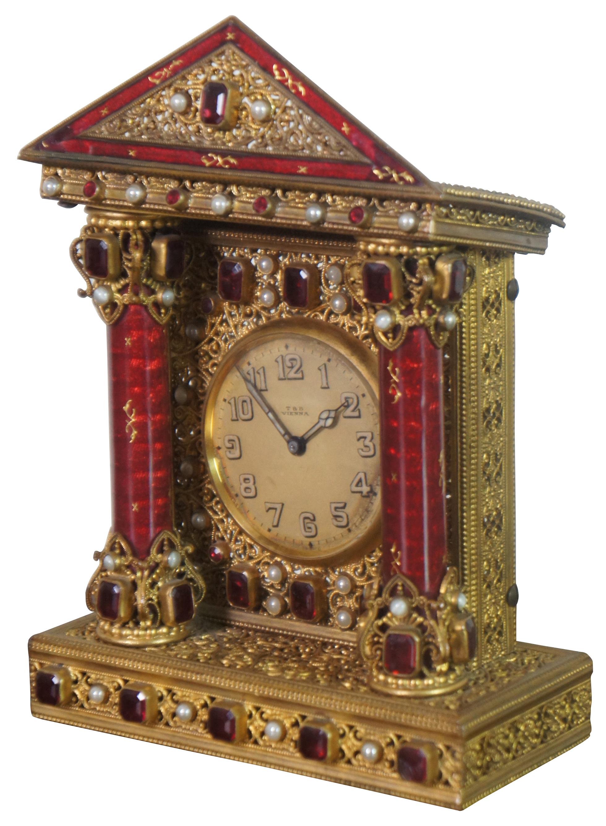 Early 20th century French table clock styled as a neoclassical building crafted of gilded filigree studded with gemstones and pearls, featuring two columns of semi-translucent red enamel columns supporting a triangular pediment. Dial marked T&B