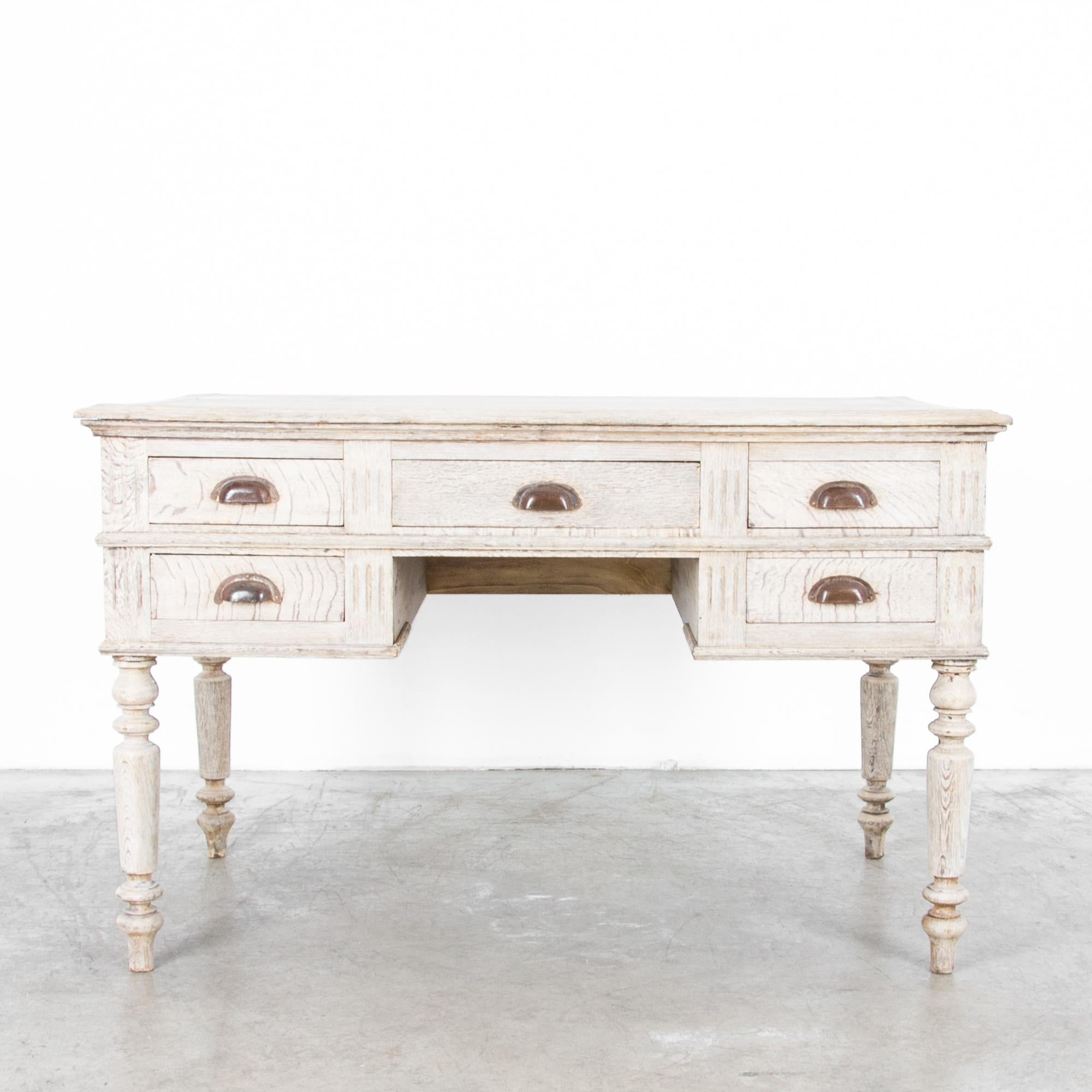 A oak desk from France, circa 1900, with five spacious drawers. A restrained neoclassical silhouette features tuned legs with a gentle taper and a tiered table ledge, while each drawer sports a metal drawer pull in a weathered bronze tone.