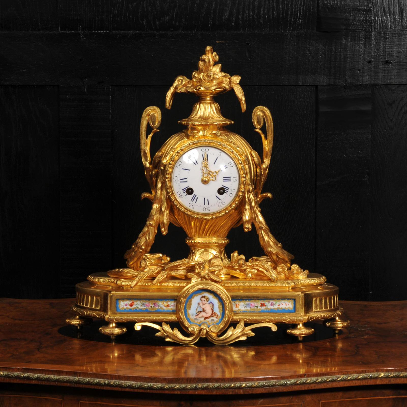 An exquisite antique French ormolu clock mounted with Sevres style porcelain. Made in finely gilded bronze, sculptured as a large gadrooned and handled urn, draped with acanthus garlands and decorated with flowers and foliage. The square base with