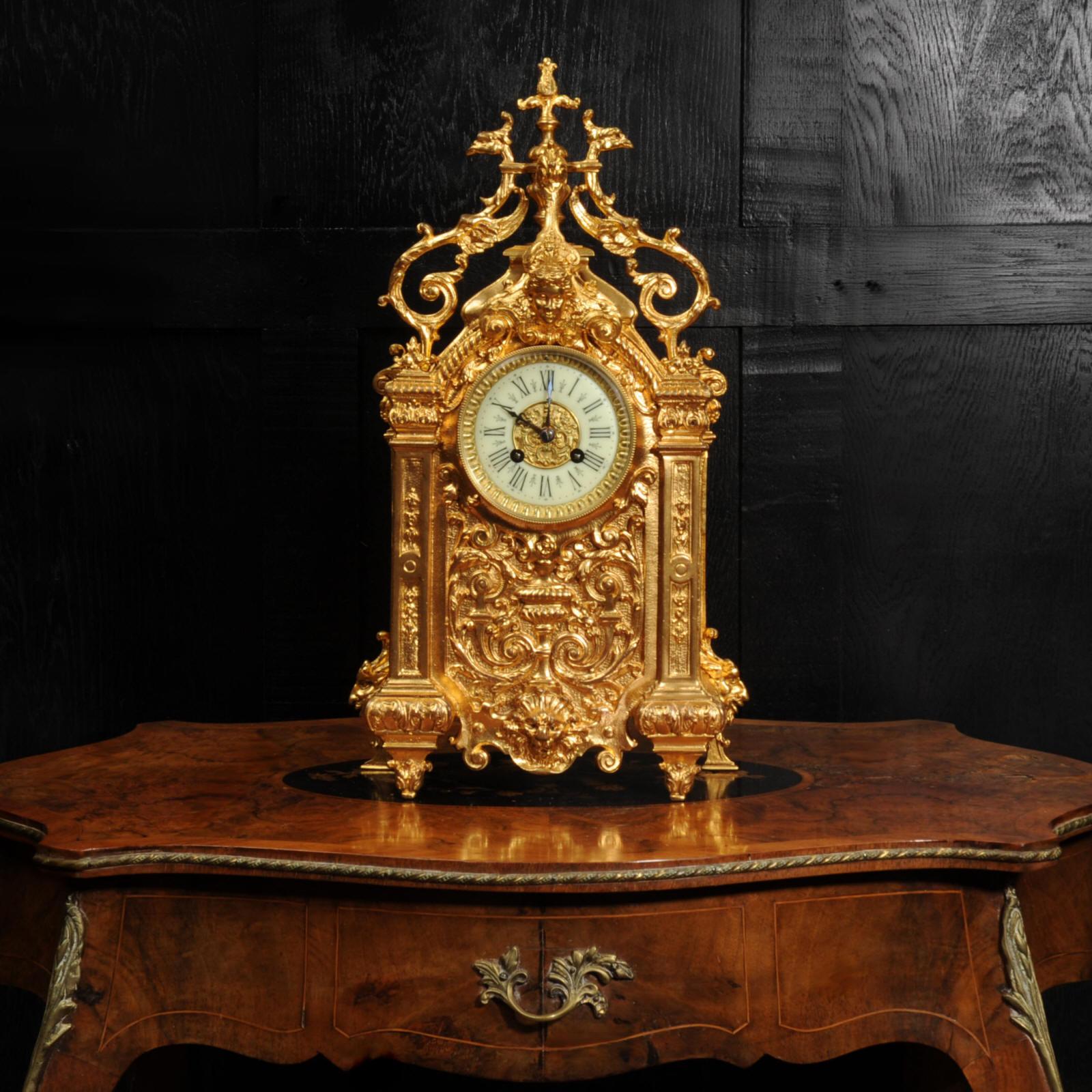 A beautiful antique French clock in finely gilded bronze. It is Baroque in style with a goddess mask, a lions mask and mythical creatures. The front is architecturally designed with two Corinthian columns with a panel of an urn sounded by scrolling