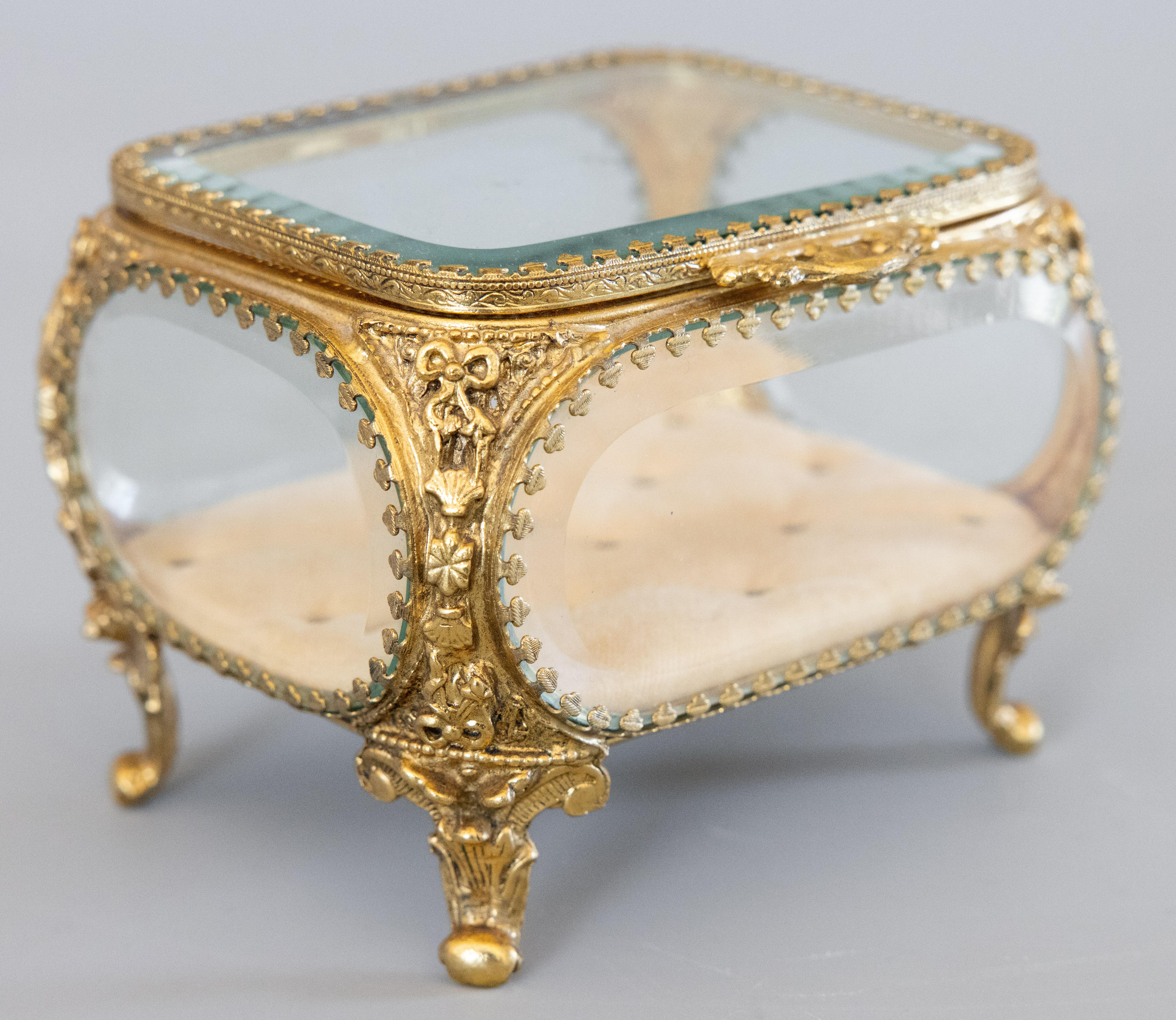 A gorgeous antique early 20th-Century french ormolu glass footed jewelry trinket box or dresser box with hinged lid and lovely shell shaped handle. This fine quality box has the original tufted velvet lining, thick beveled glass, intricate gilt