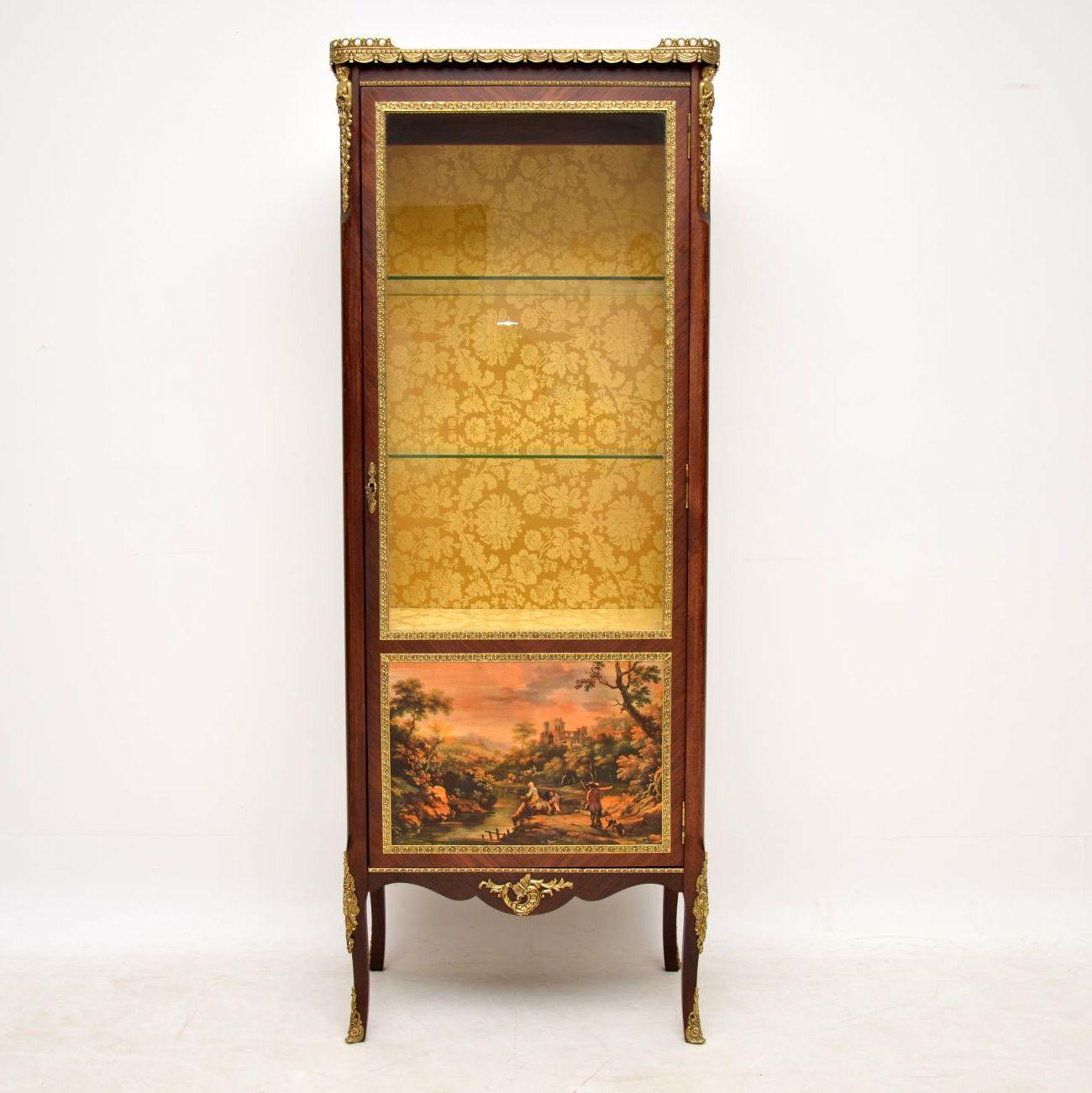 Antique French style mahogany display cabinet with very detailed classical gilt bronze mounts. The top gallery has pierced work with swags below. The inside of this cabinet has gold embossed fabric and glass shelves, plus more storage space below.