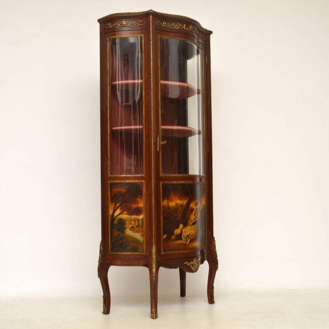 Antique French style display cabinet with a serpentine shaped front in mahogany and with decorative scenes on the bottom panels. There are also a lot of gilt bronze mounts all over this cabinet. It's in good original condition and still has the