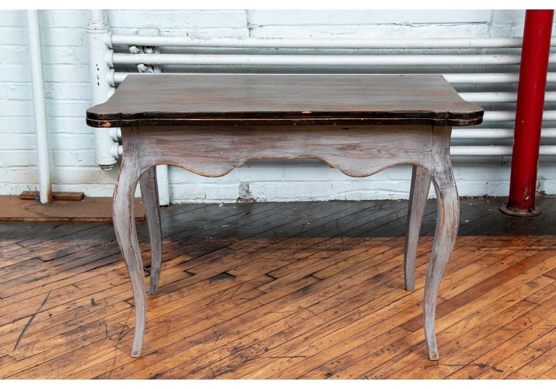 Une belle table avec un tablier serpentin et des pieds cabriole dans une peinture grise plus récente et un plateau teinté foncé dans une finition antiquaire. Le plateau à charnière est peint en brun foncé avec des coins façonnés. L'un des pieds sert