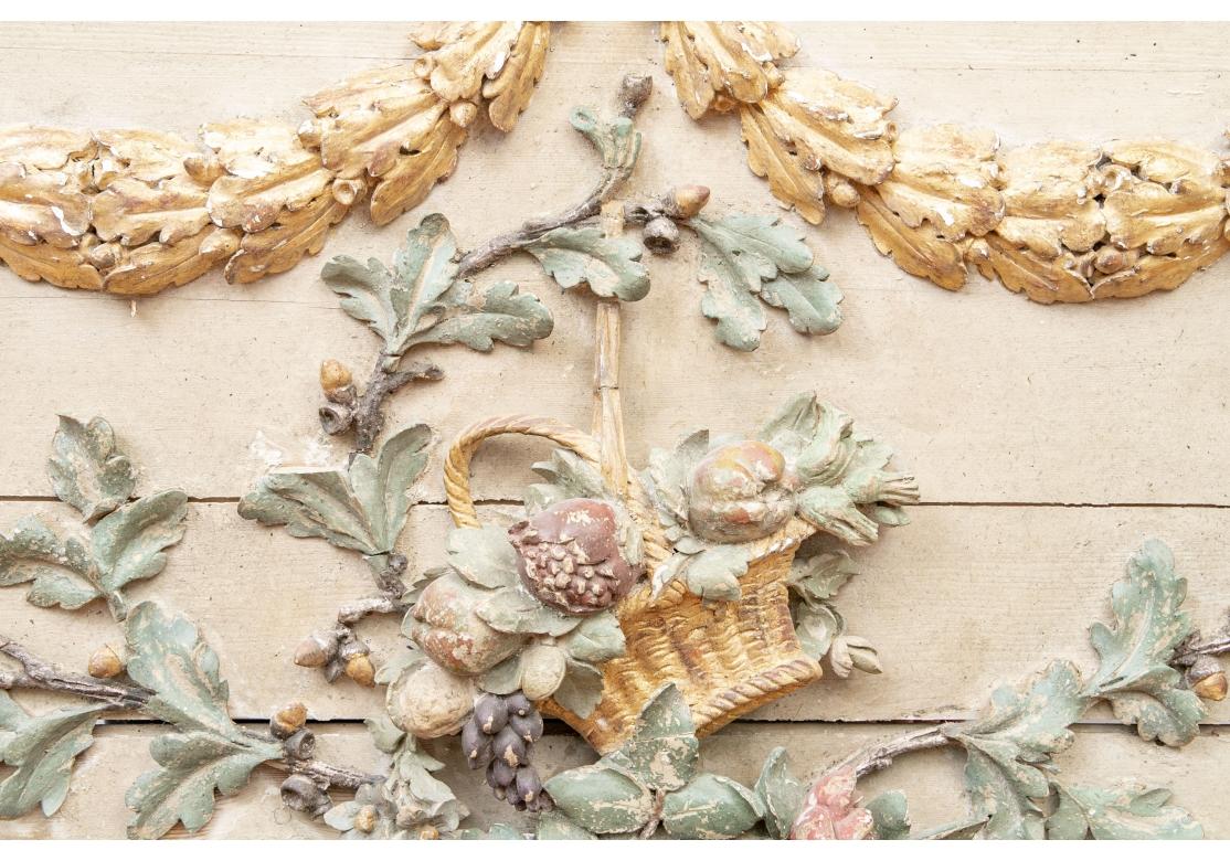 Originally purchased from Hollyhock in West Hollywood, California. A large panel in ecru paint with carved painted and gilt high relief decoration- a gilt bow and swags at the top over a basket of fruits with a garden rake. All on a tall curved
