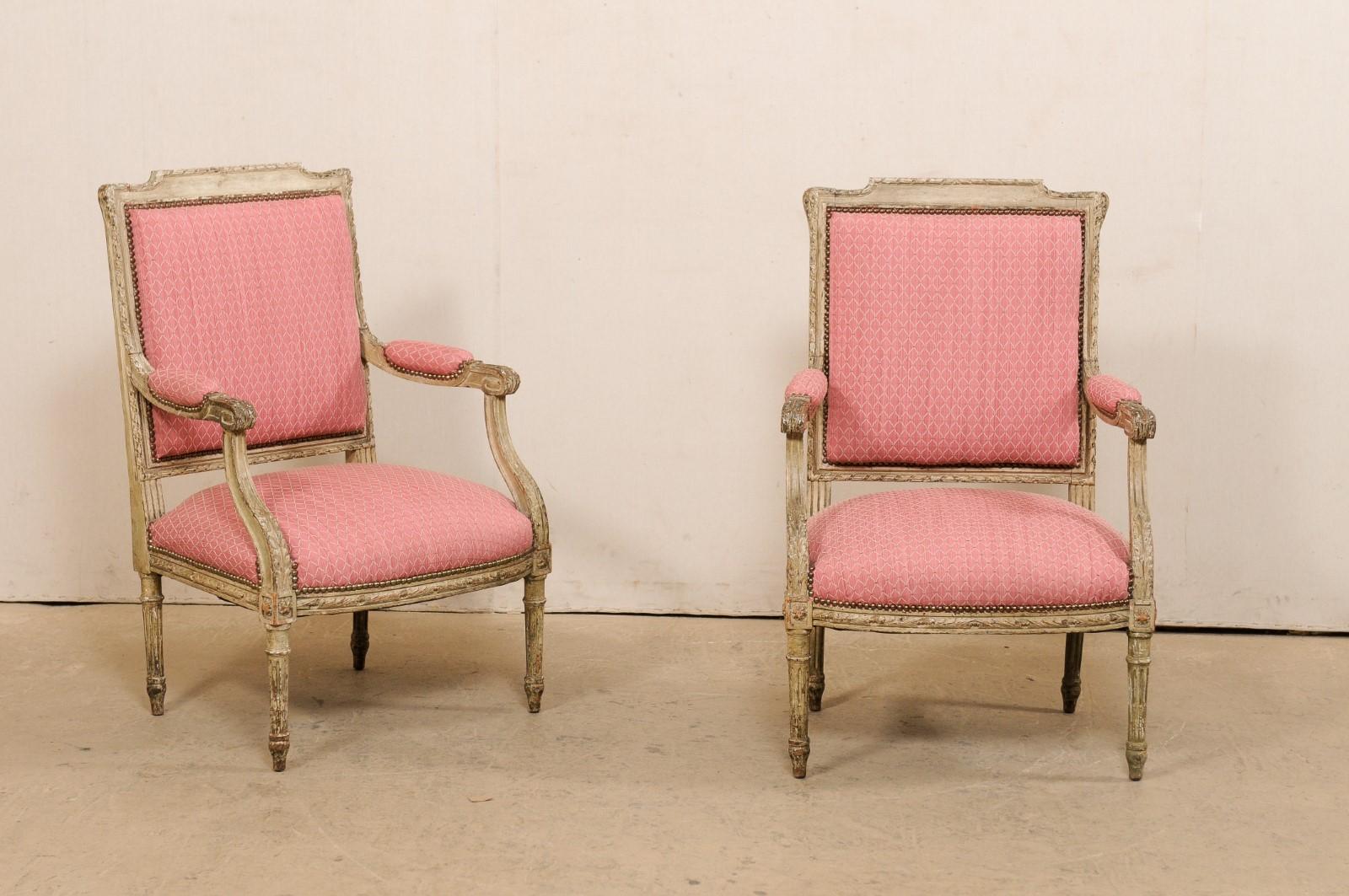 A French pair of Louis XV style, carved-wood fauteuil armchairs from the early 20th century. This antique pair of armchairs from France have upholstered backs framed within a beautifully carved wood frame edged in ribbon trim. The wooden arms, with