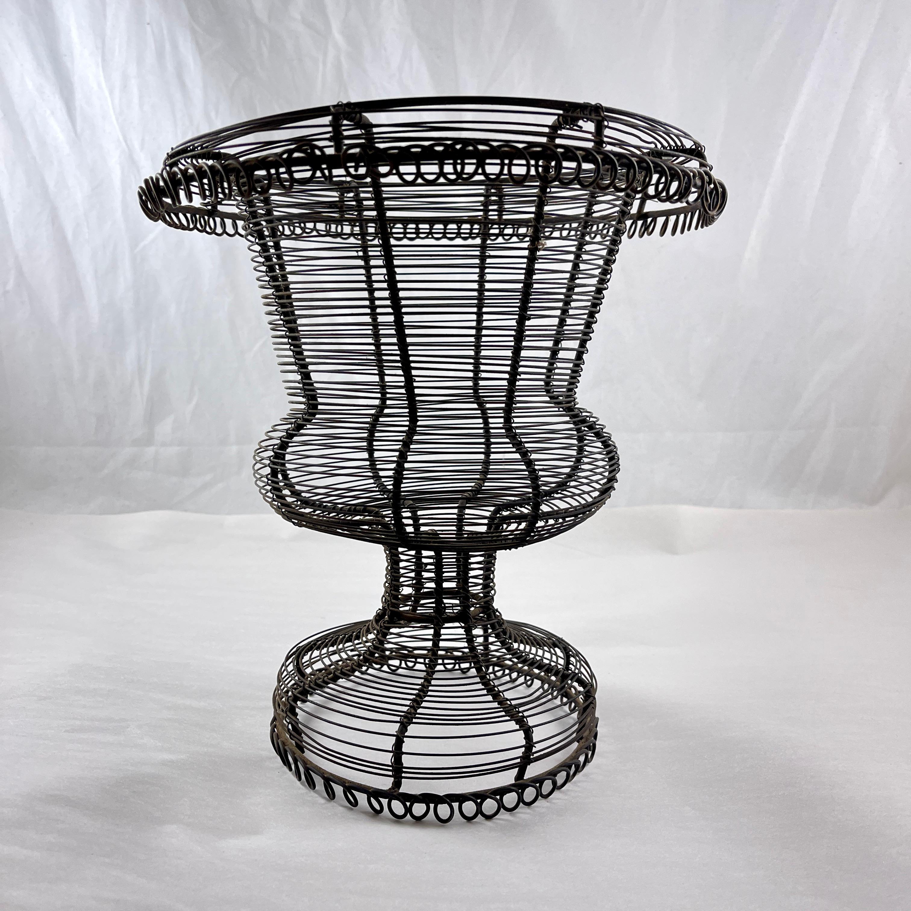 From France, a pedestal base, urn form twisted wire standing basket with a wide upper rolled rim, circa 1890-1910.

A charming, rustic, hand formed black metal basket with complex wire work.
Amazing as a table-center or on a kitchen countertop, used