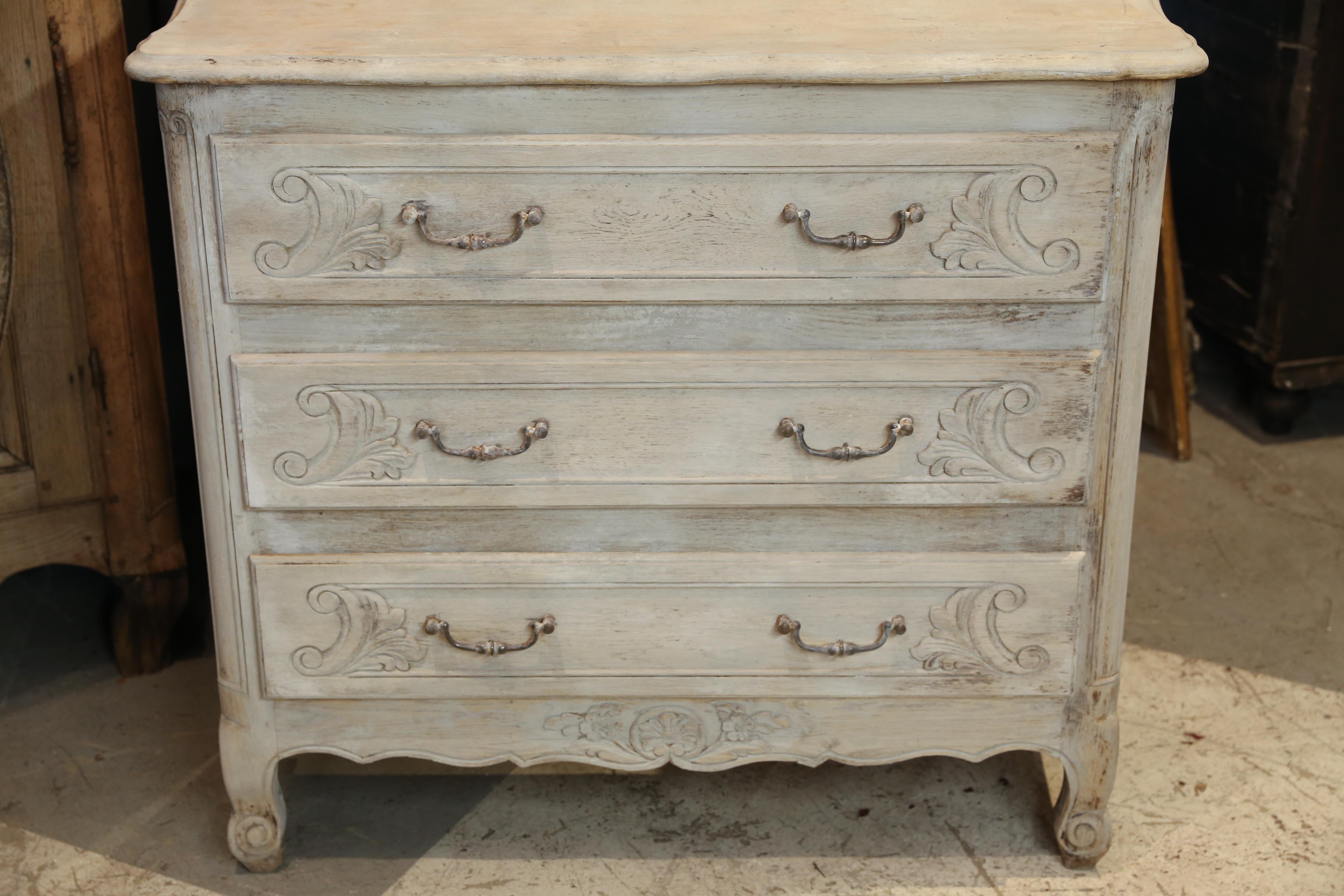 This charming and petite antique French chest of drawers features beautifully hand carved drawer fronts with iron hardware. The piece has been hand-washed with a pickled finish to brighten the wood and bring out the grain. The top of the piece has a