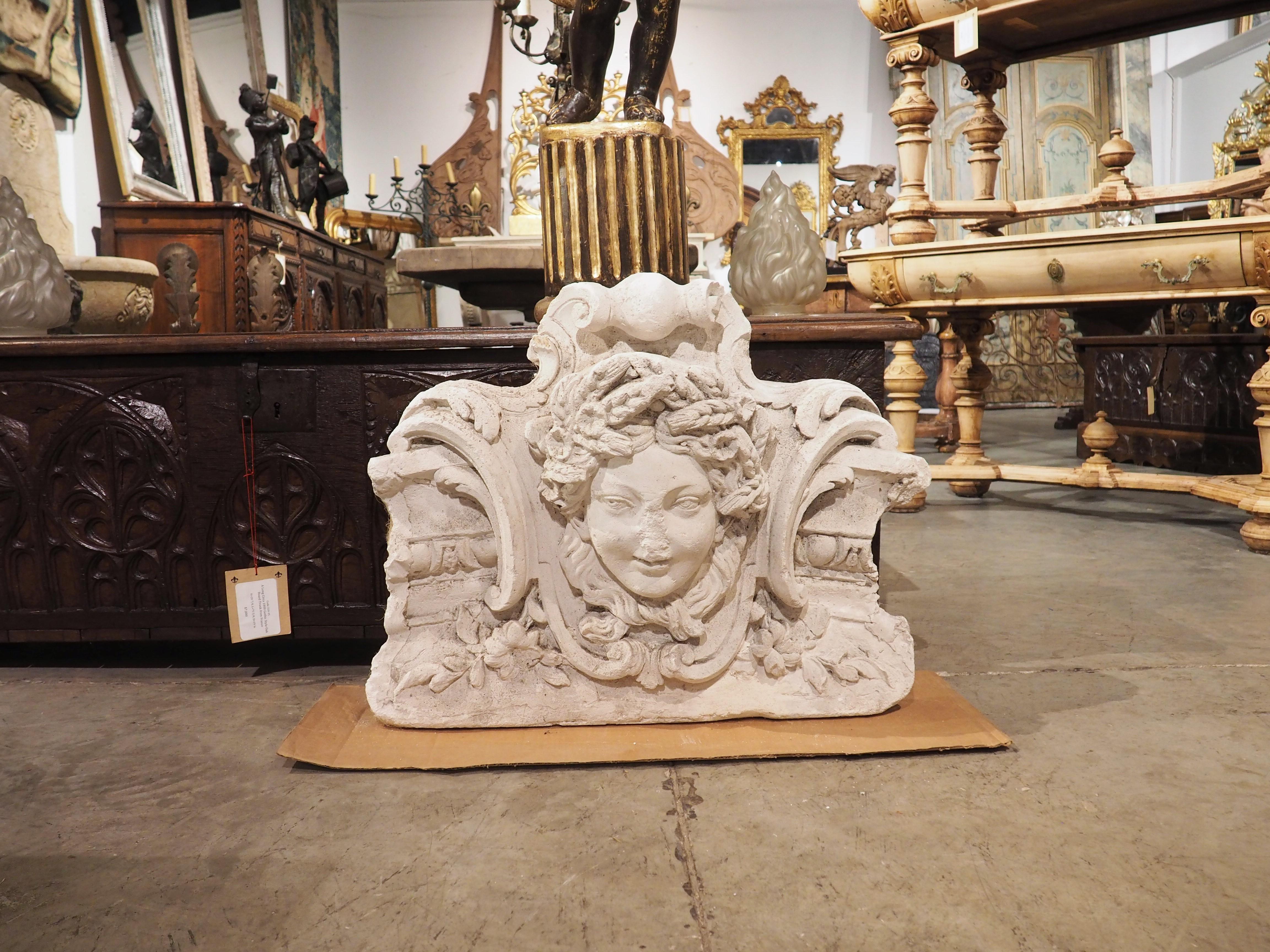 A beautiful plaster architectural from the late 1800s, depicting a female mascaron surmounting a highly scrolled coat of arms shield. The young woman has a floral headpiece atop her head, echoing a flowering margent that emanates from the base of