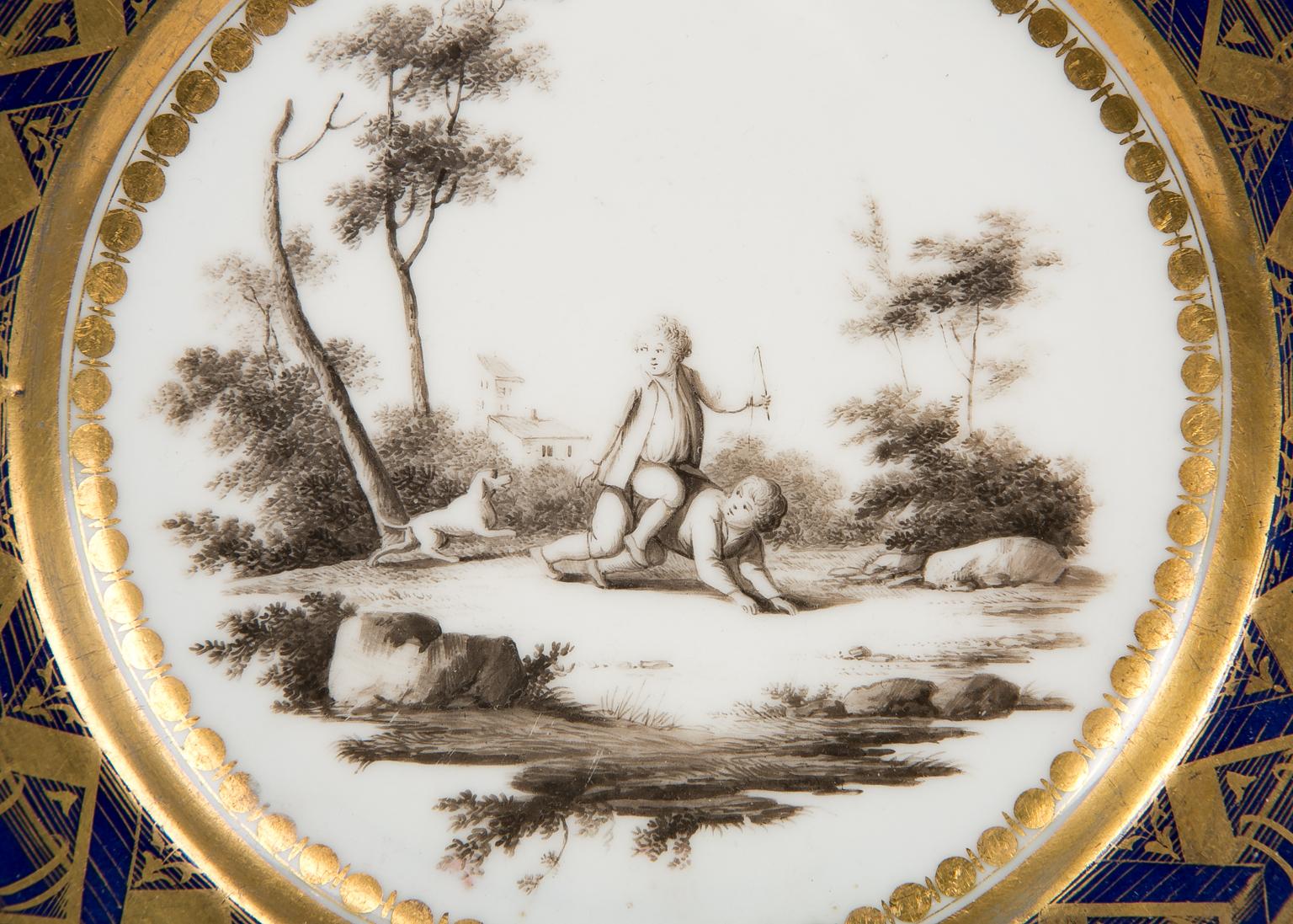 Made circa 1785, this plate features a deep cobalt blue border that is beautifully gilded with a dramatic geometric pattern. The burnished gilt is a hallmark of Dagoty porcelains. 
Inside the dramatic border is a wonderful scene of children playing