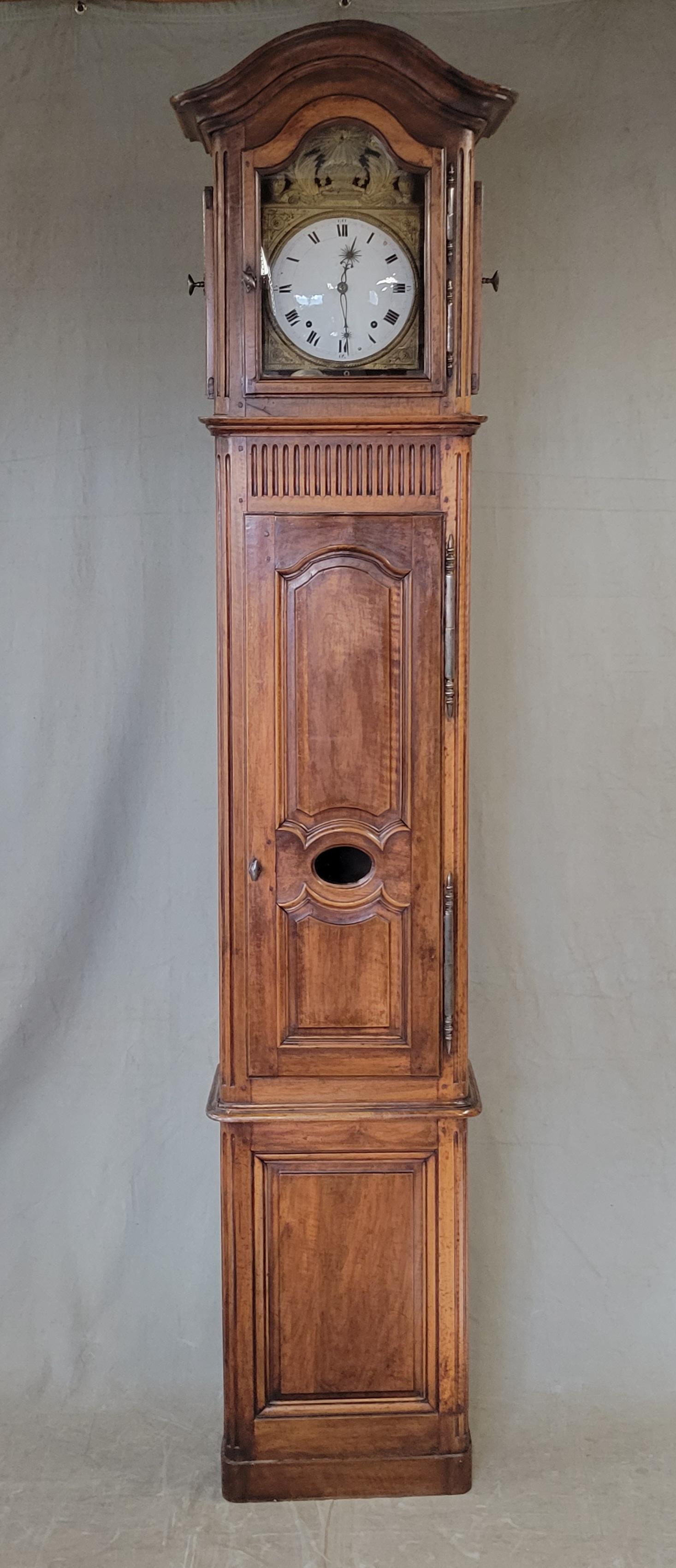 A stately antique French Provincial walnut tall case clock with an enamel face and embossed brass surround. Charming blue print wallpaper on the inside of the top clock case portion. Side door open which enables winding of the clock. Clock winding